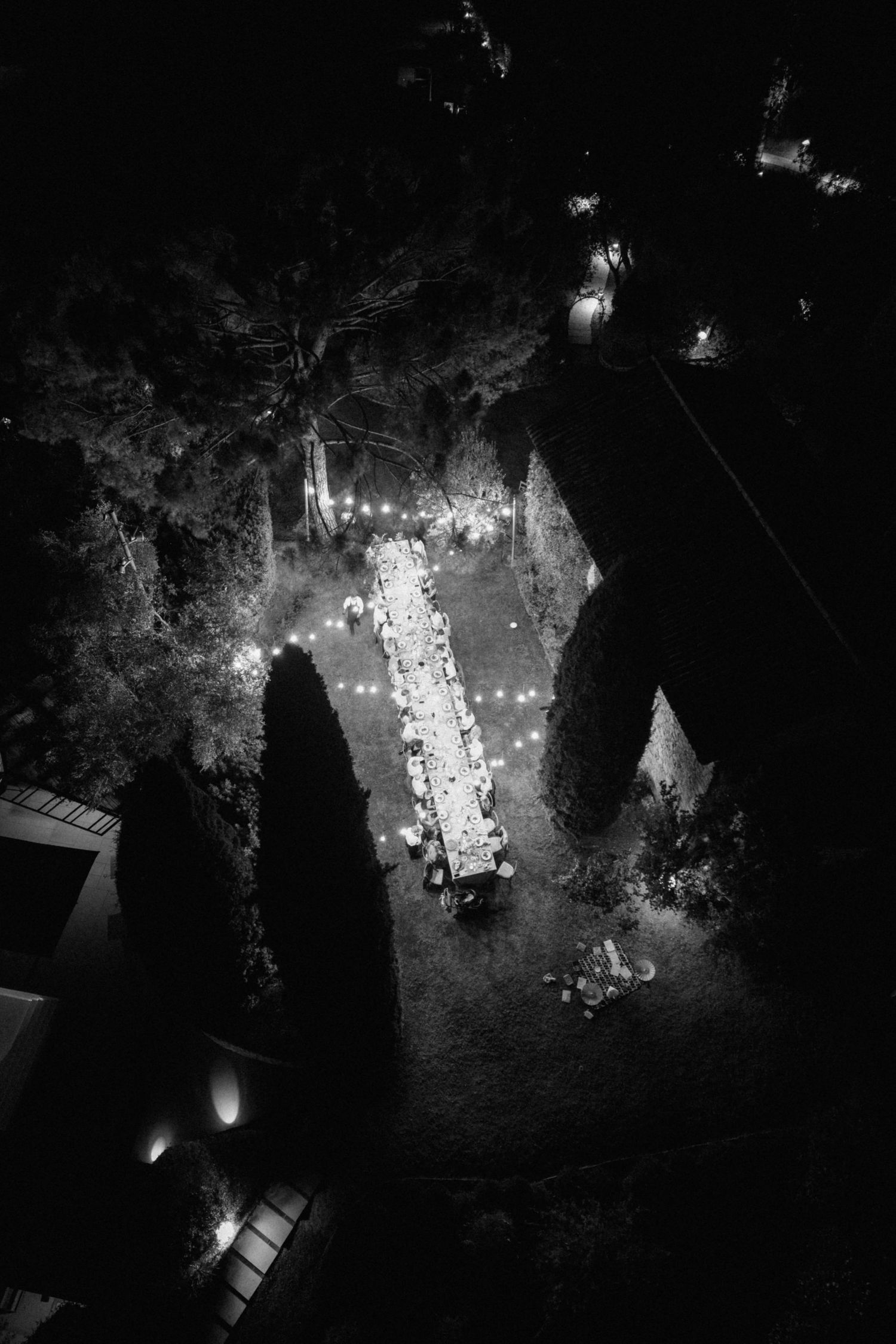 Aerial black and white night view of an outdoor wedding reception table with string lights in a garden.