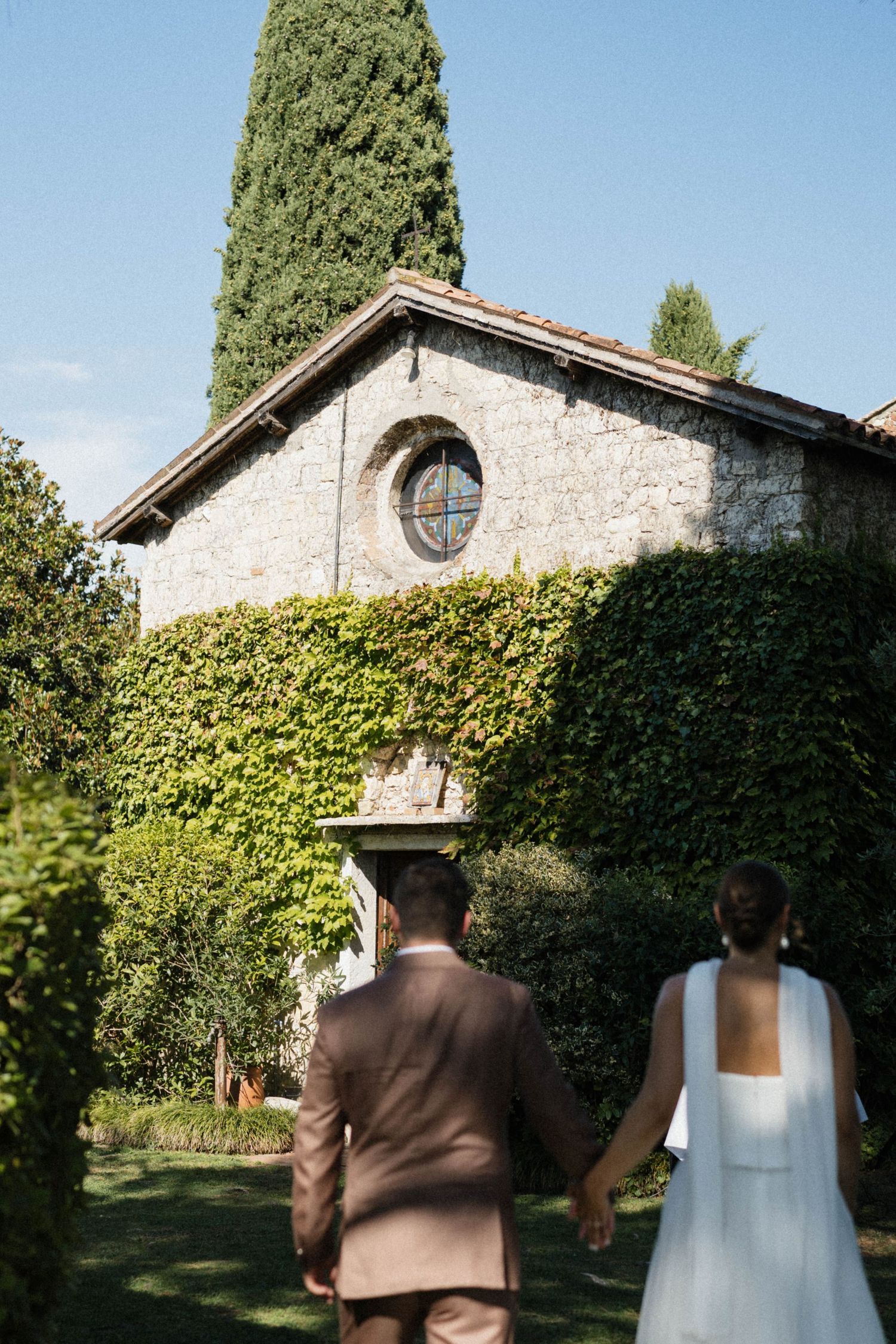 Bride and groom hold hands walking toward an ivy-covered stone chapel with a cypress tree.