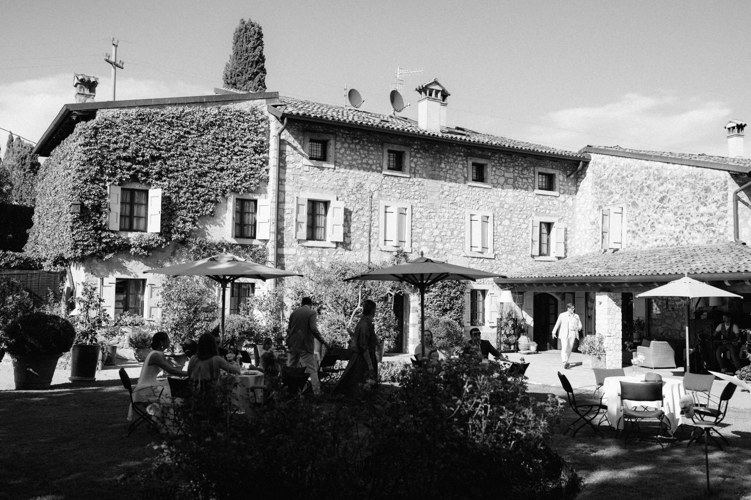 Black and white photo of a rustic ivy-covered stone farmhouse with outdoor seating and umbrellas in a garden setting.