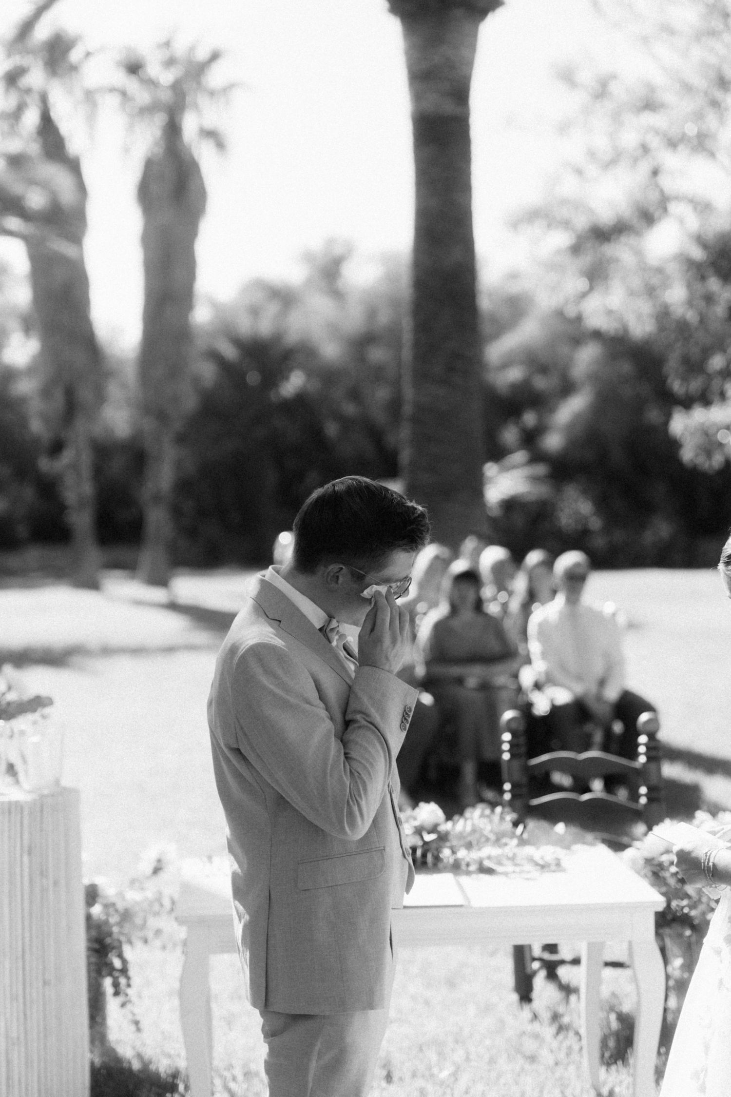Emotional groom wiping tears during outdoor wedding ceremony surrounded by palm trees.