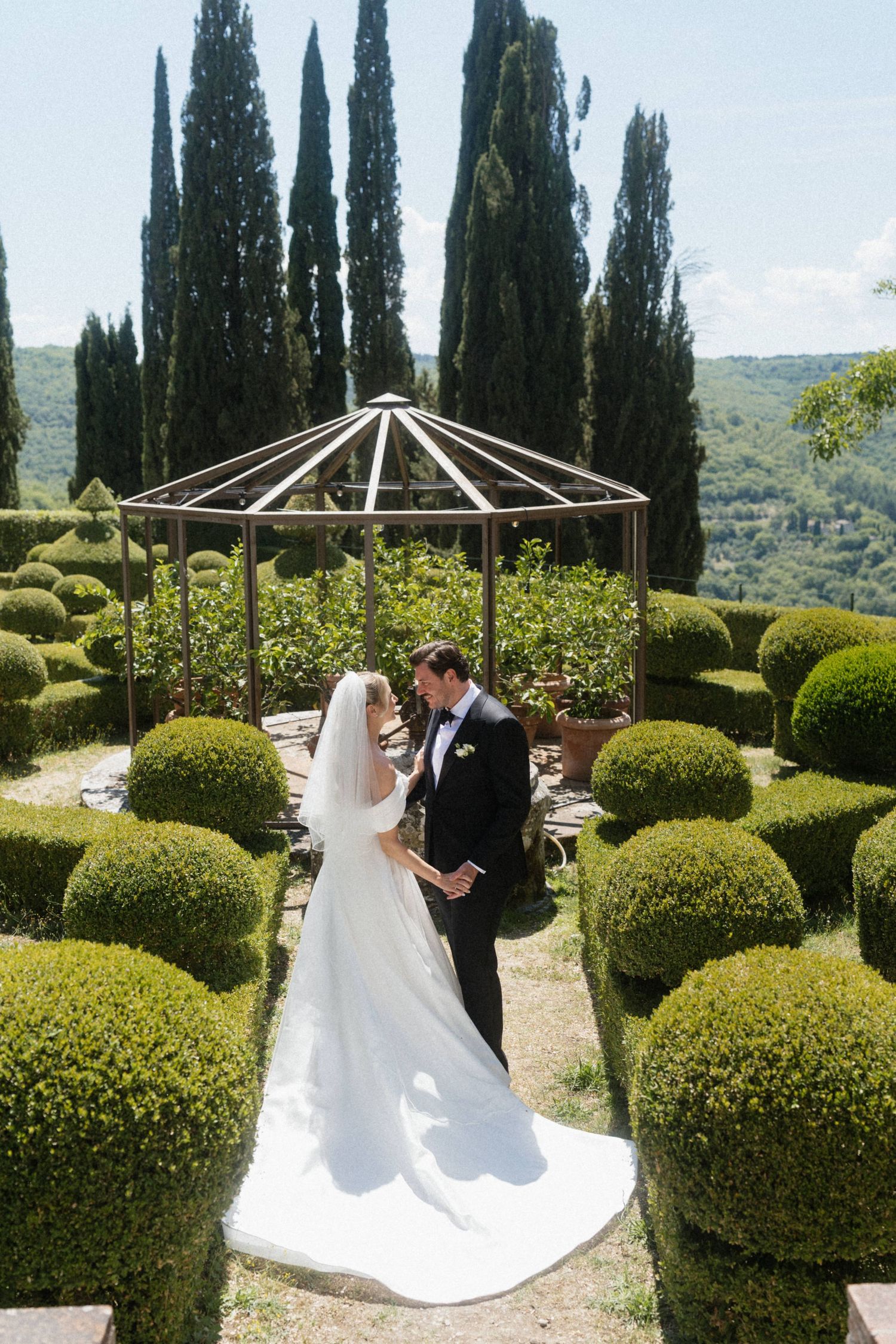 Bride and groom share romantic moment near gazebo surrounded by Italian cypress trees and topiary garden.