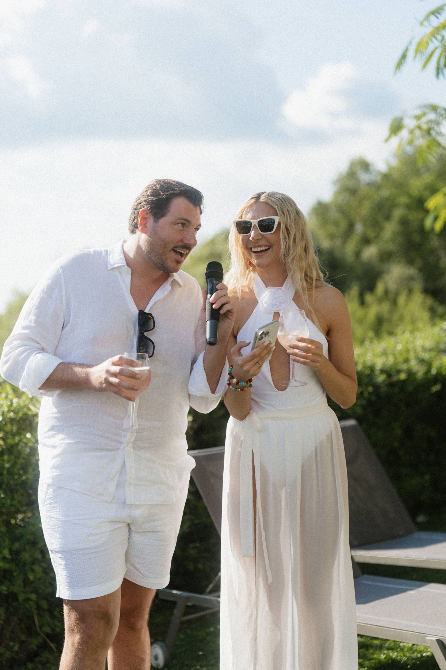 Smiling couple in white hosting a wedding event outdoors, sharing a microphone under blue skies.