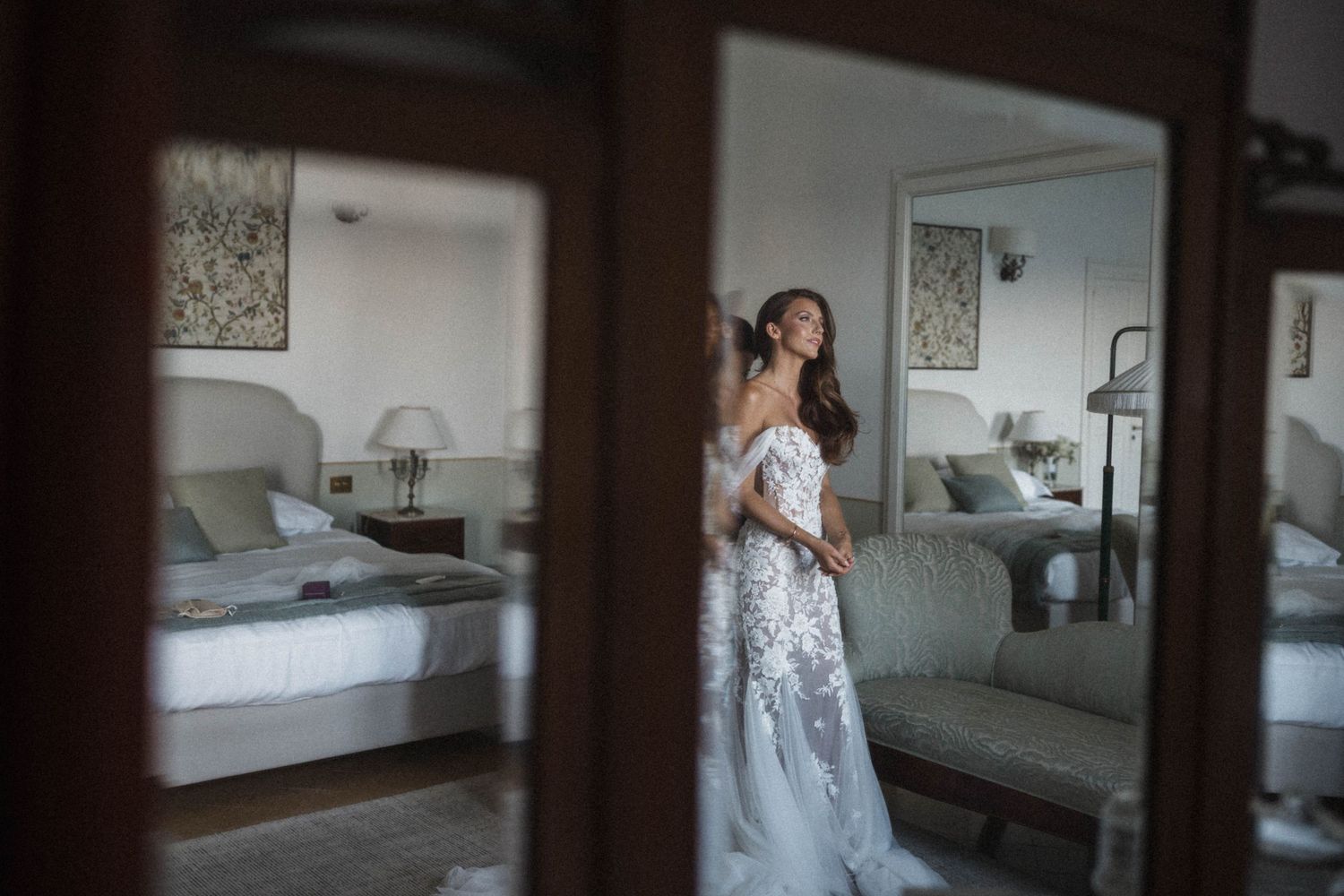 Bride in lace wedding gown preparing in elegant hotel room, reflected in mirror.