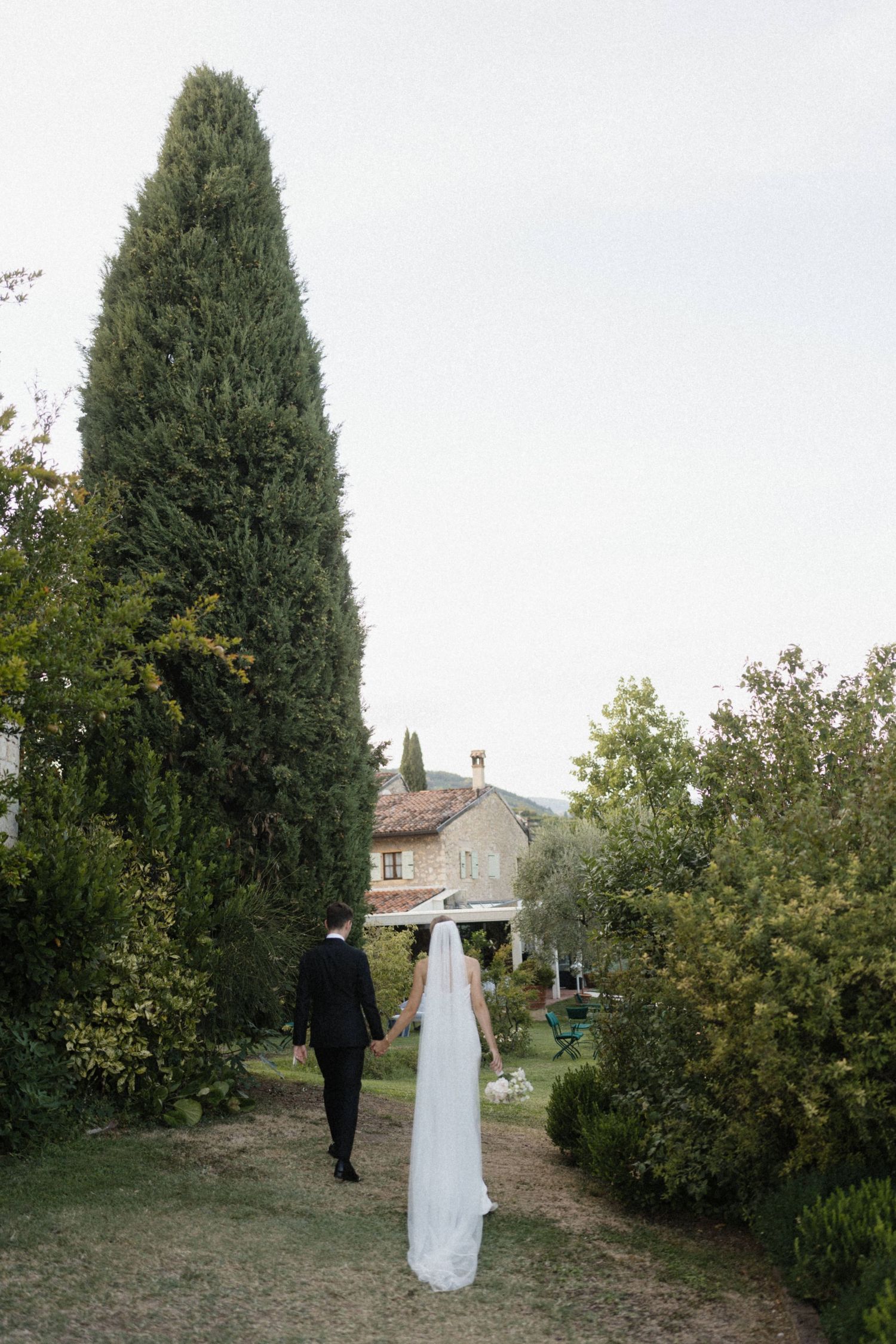 Bride and groom walking hand in hand through a lush garden with a cypress tree and stone villa in Tuscany.