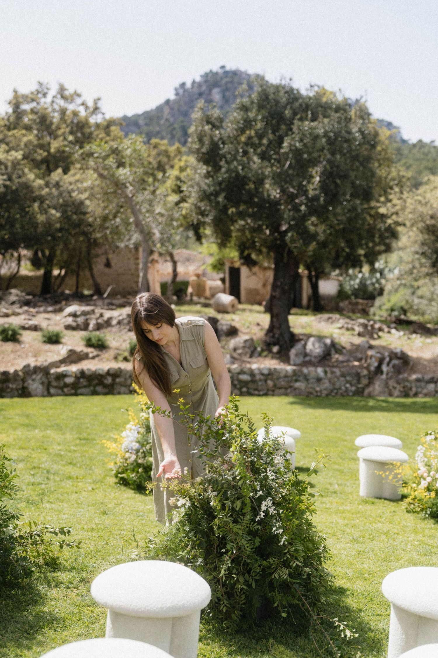 A woman in a grey dress arranges floral decorations on a sunny outdoor wedding ceremony setup with lush greenery.