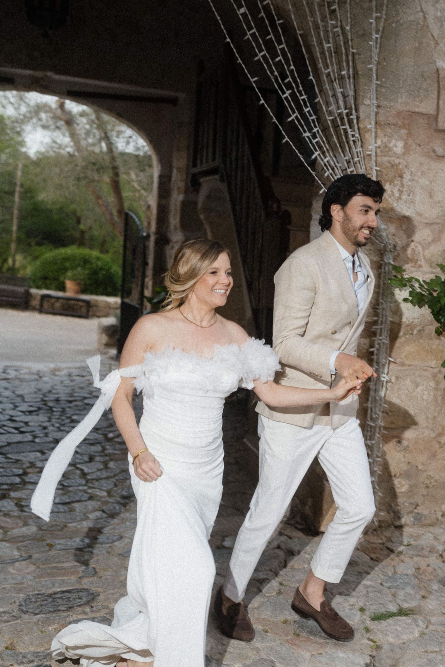 Bride and groom laughing while running through cobblestone archway at rustic wedding venue.
