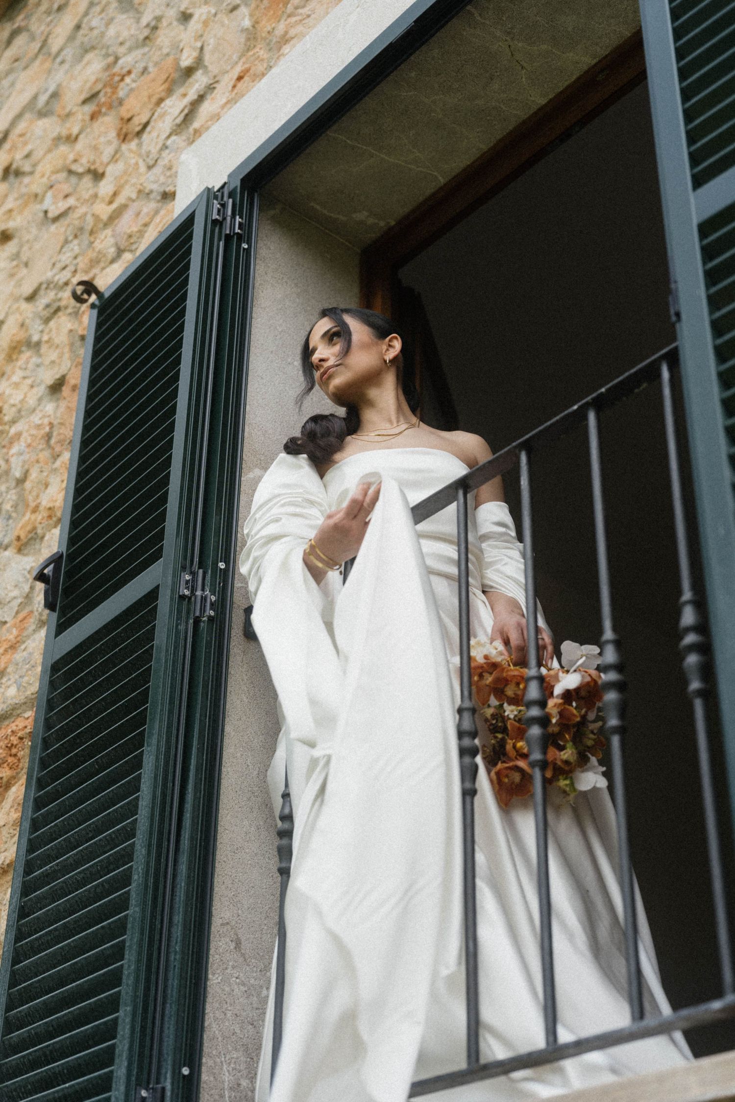 Elegant bride in off-shoulder white gown holding autumn bouquet on iron balcony with green shutters.