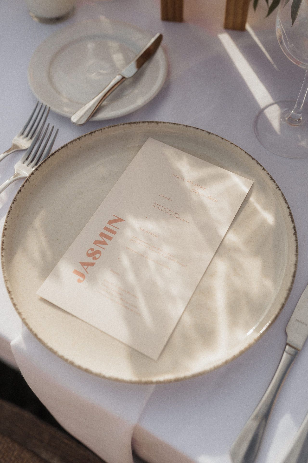Elegant wedding place setting with gold-rimmed plate, personalized menu card reading JASMIN, silverware on white tablecloth.