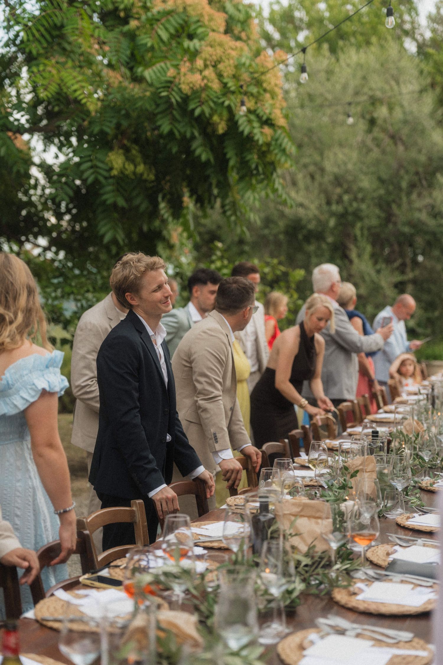 Wedding guests standing along decorated outdoor reception table with string lights and lush greenery backdrop.