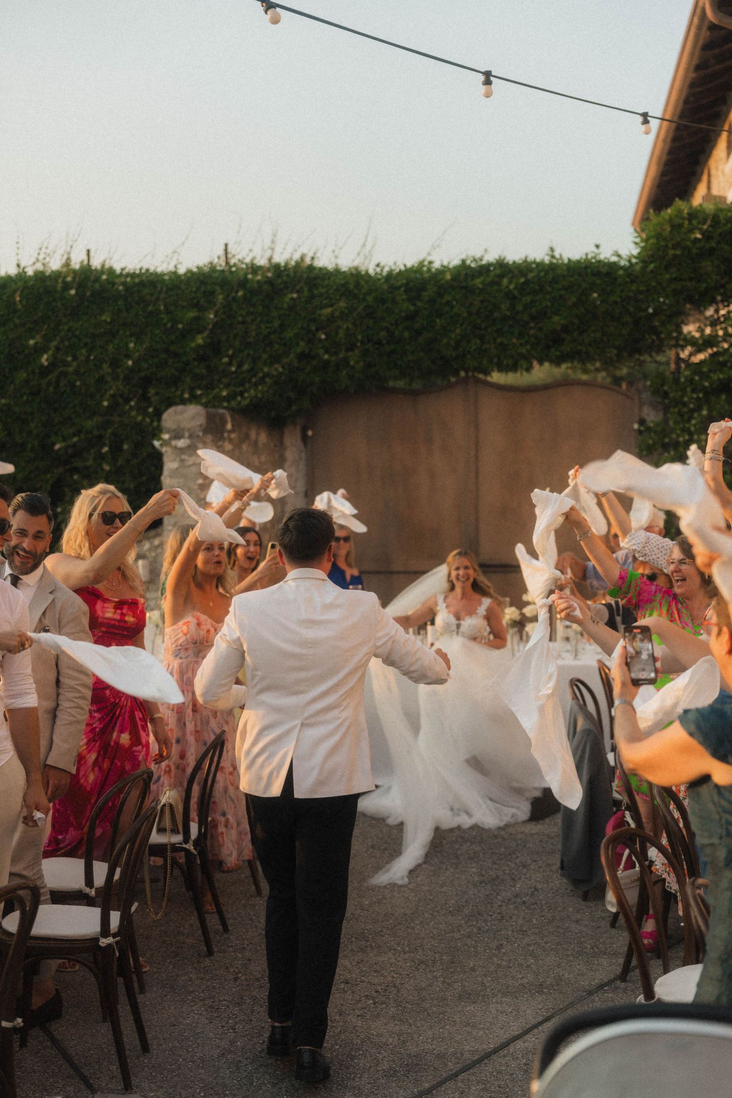 Bride and groom walk through cheering guests waving white napkins at outdoor wedding reception.