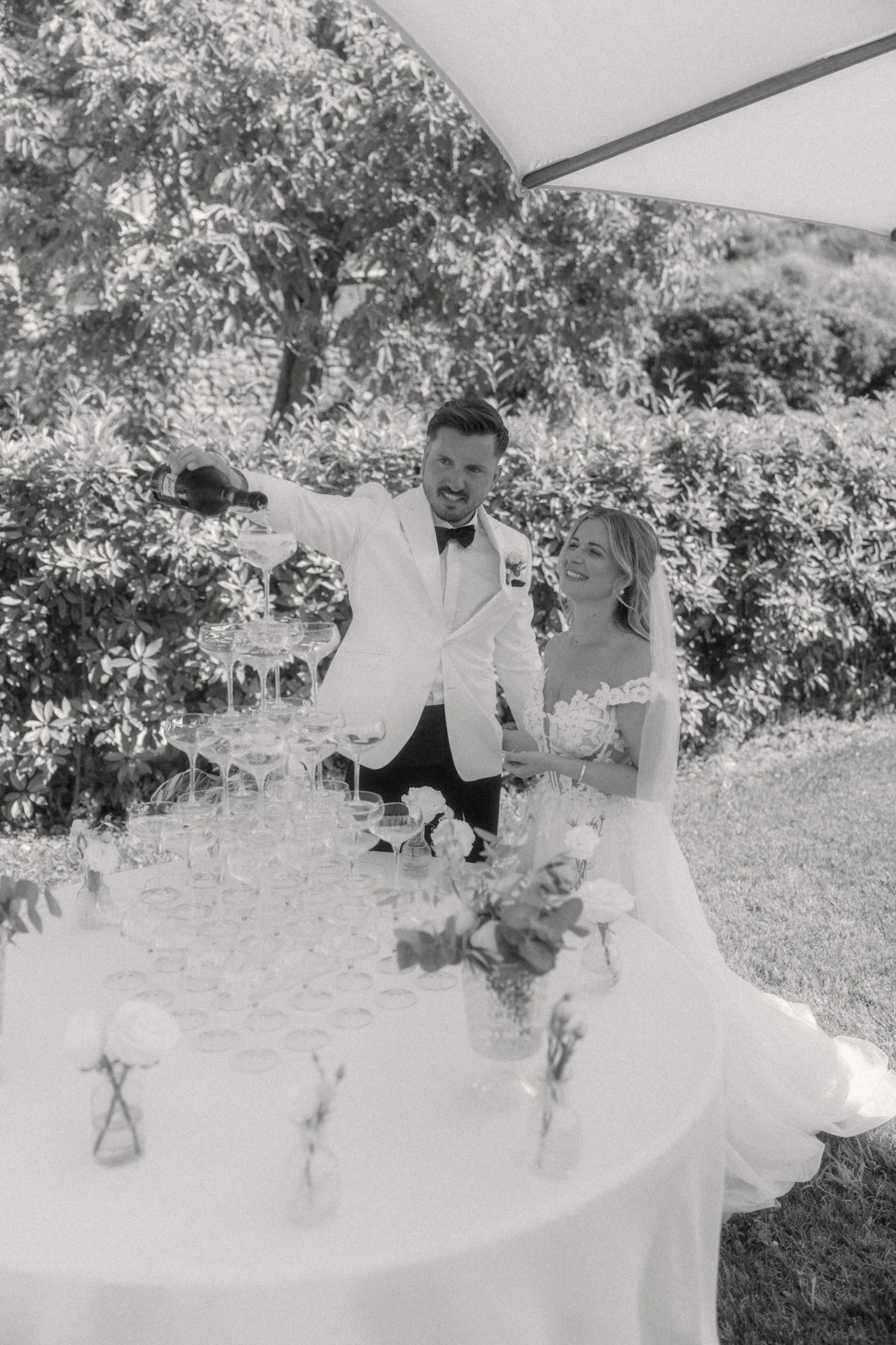Bride and groom pouring champagne tower at elegant outdoor wedding reception in black and white.