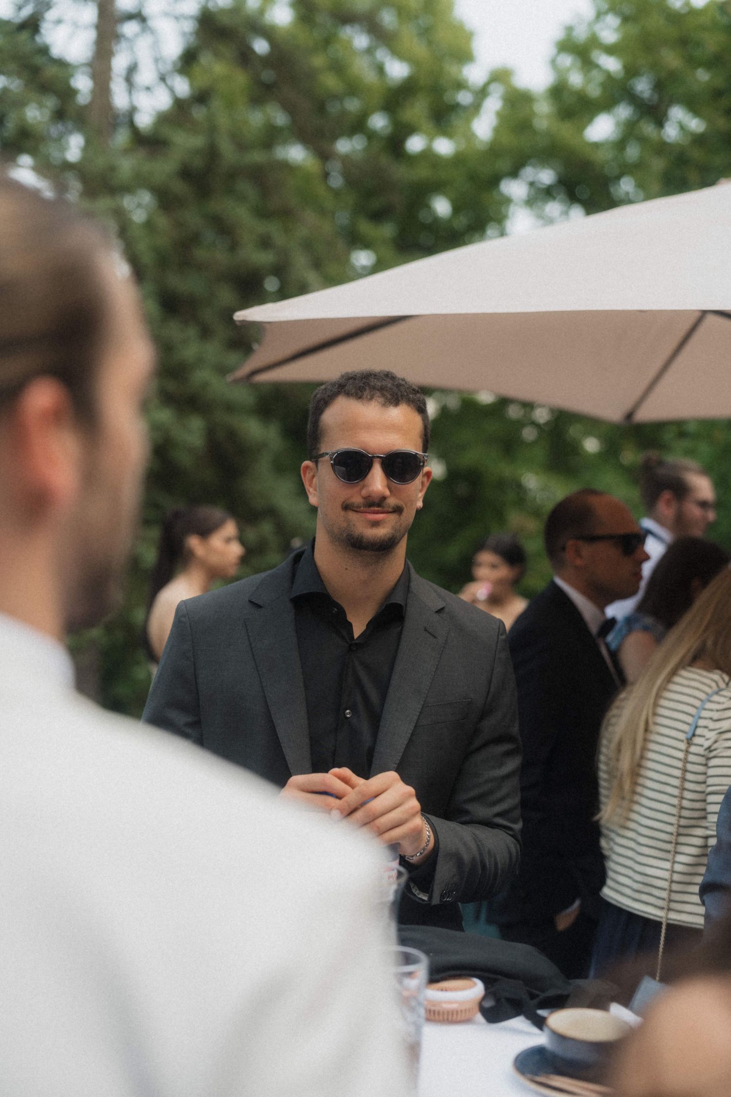 Stylish man in dark suit and sunglasses smiling at an elegant outdoor wedding reception.