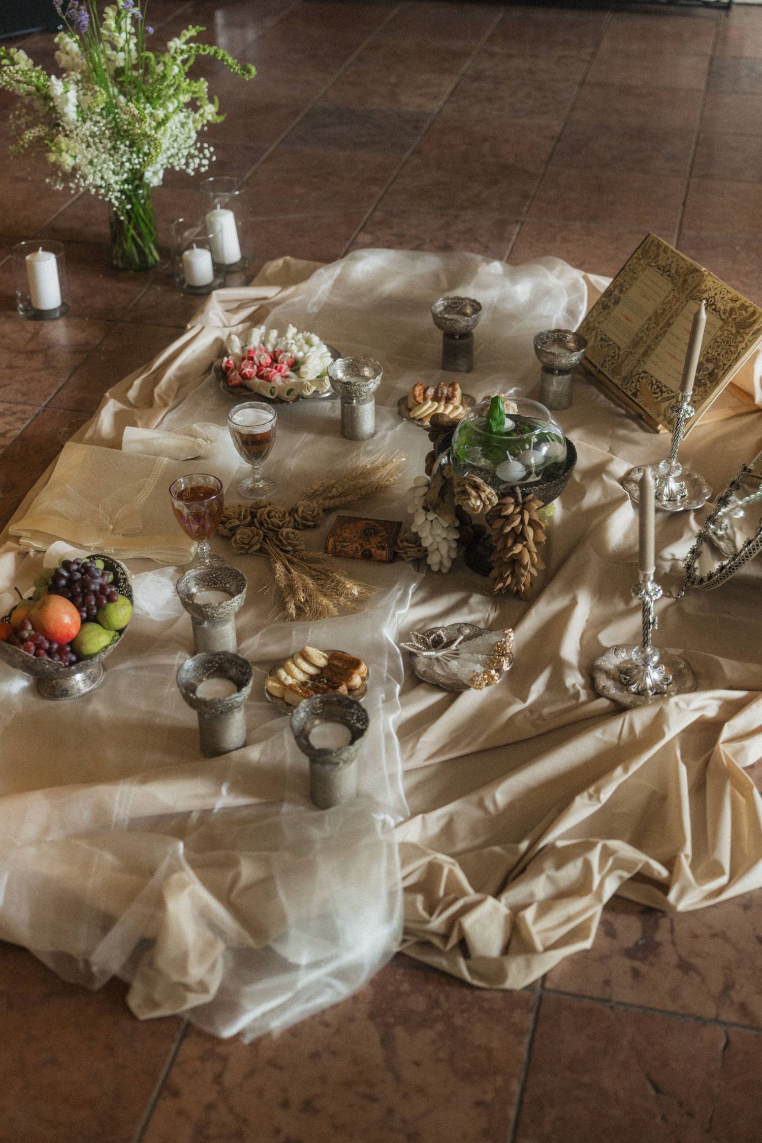 Elegant wedding ceremony floor setup with silver goblets, fruits, candles, and ornate decorations on a cream silk cloth.