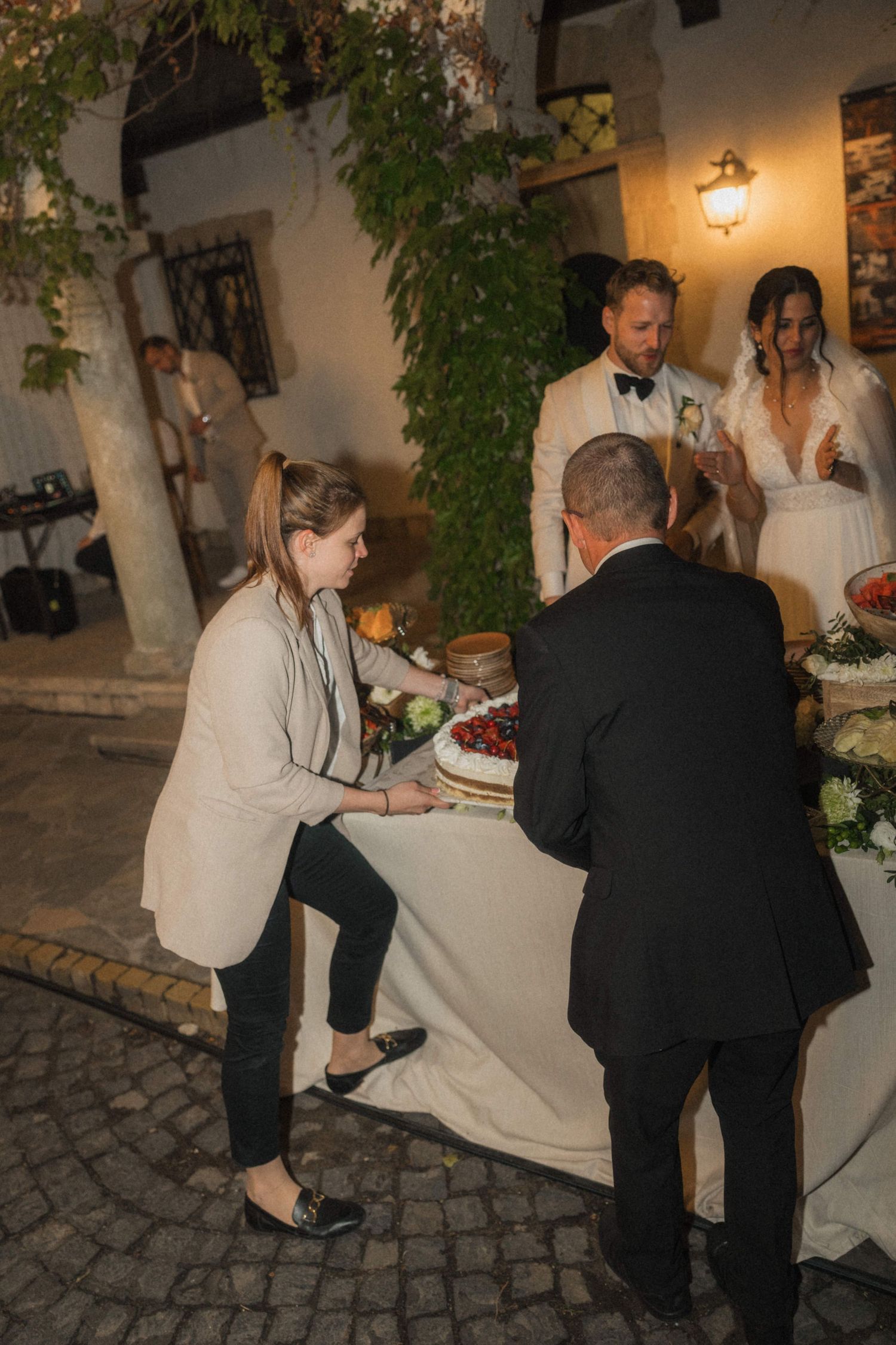 Wedding planner arranges cake table as bride and groom in white stand nearby at night.