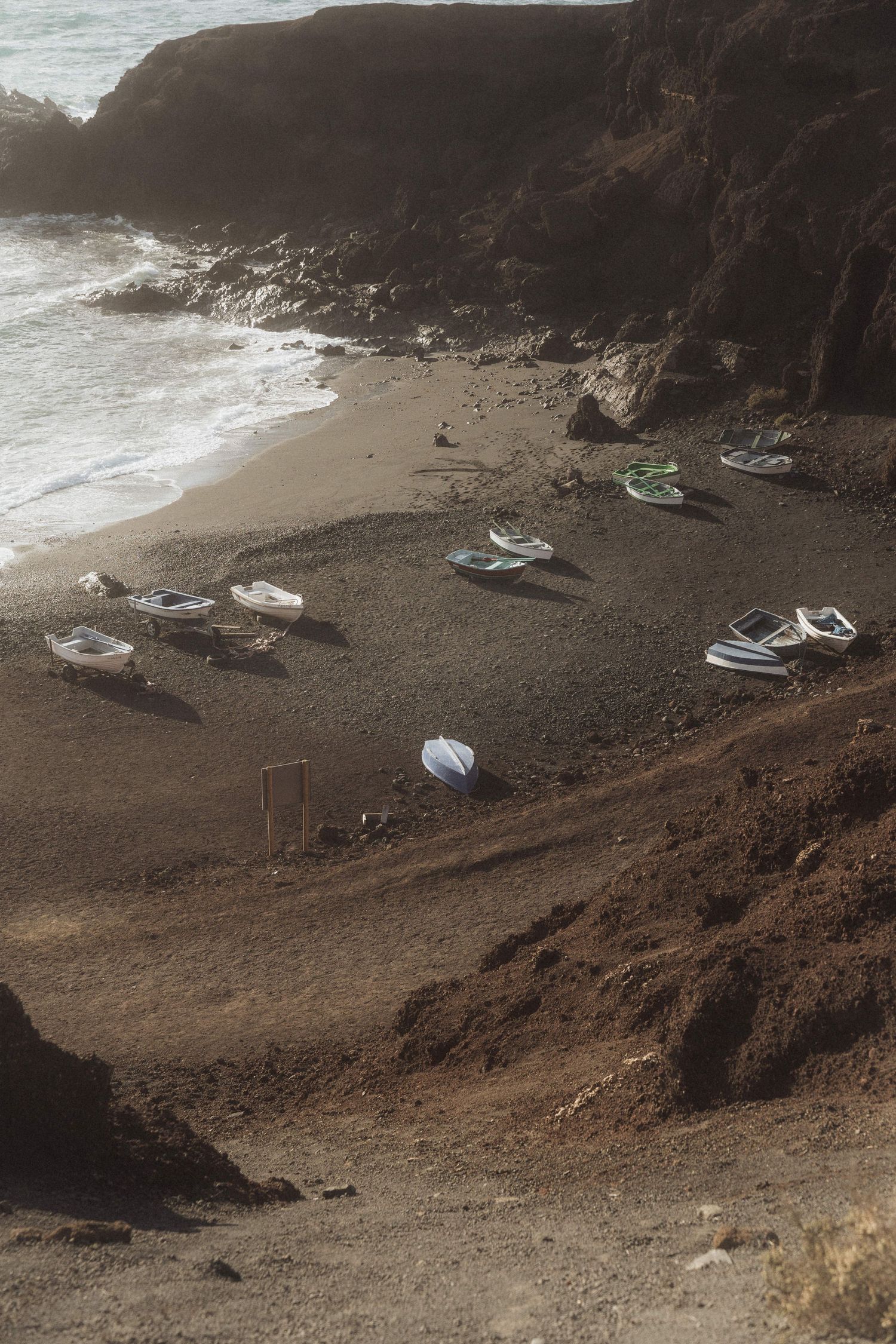 Aerial view of small fishing boats resting on a secluded dark sand beach surrounded by rocky cliffs.