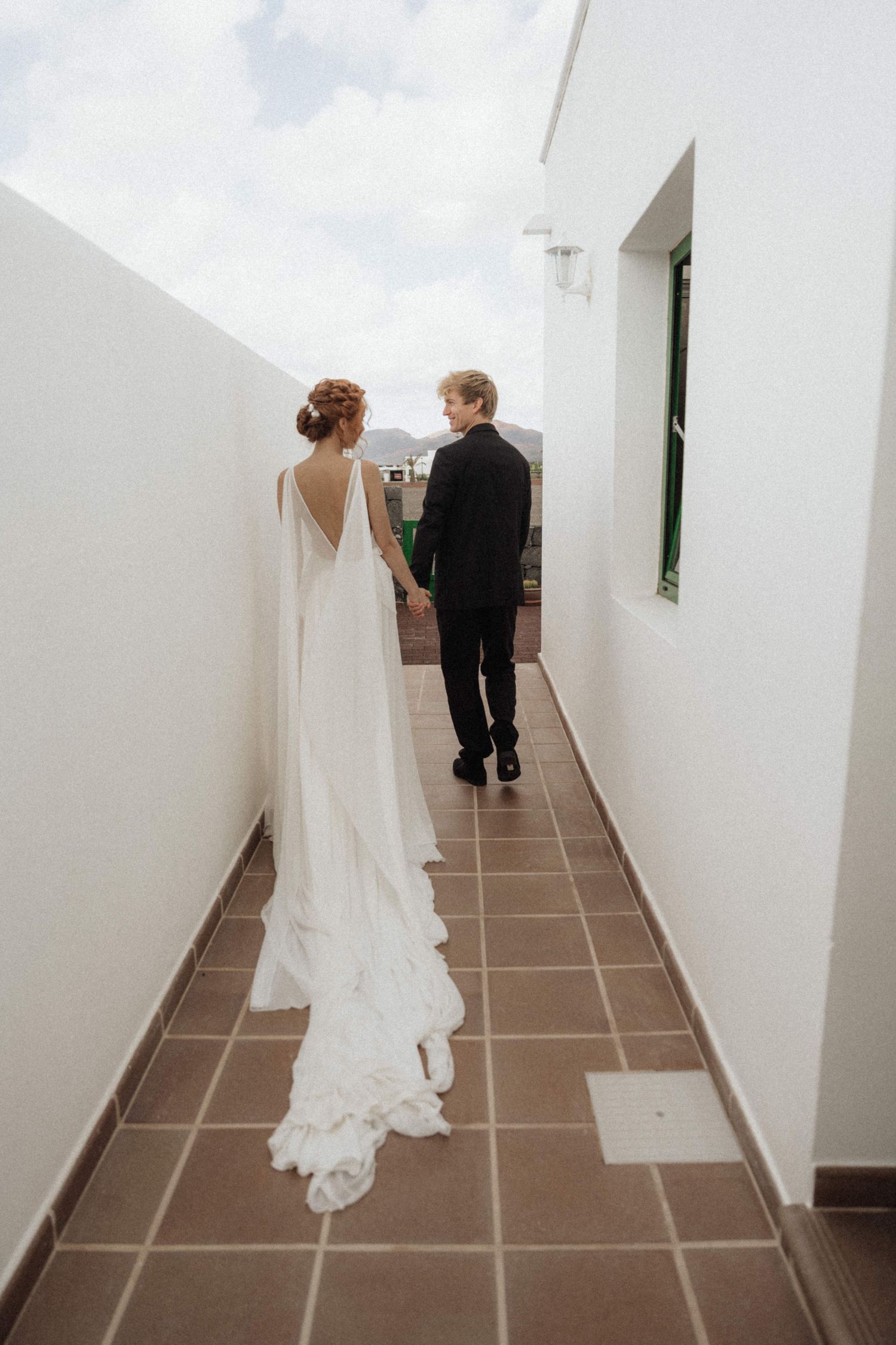 Bride in elegant white cape gown holds hands with groom in black suit walking through a narrow outdoor corridor.