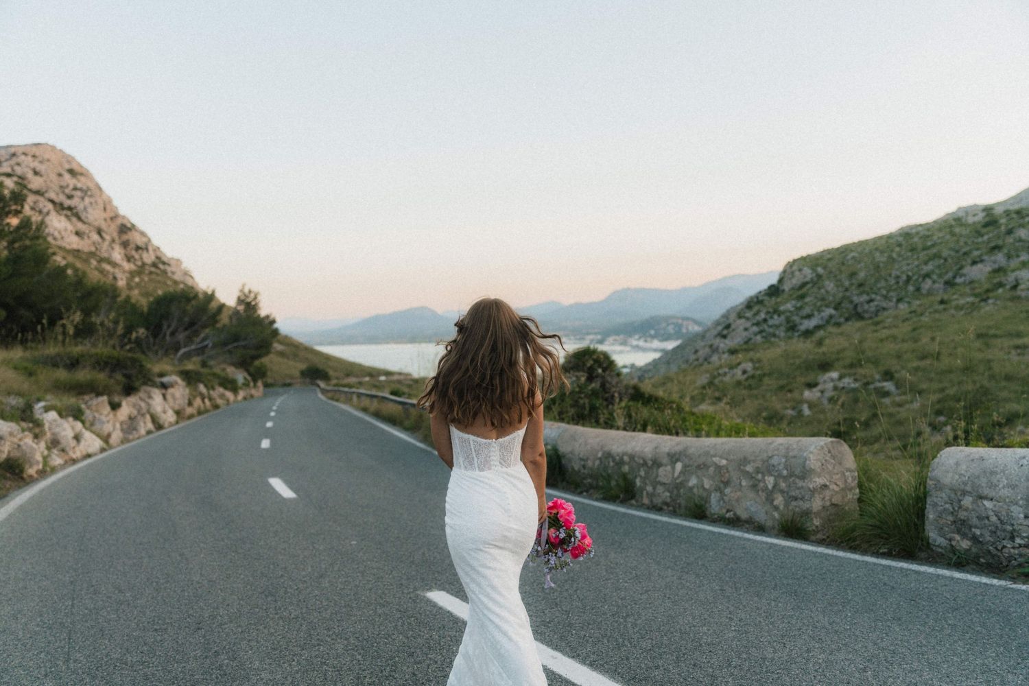 Bride in white fitted gown walking down scenic coastal road surrounded by green hills and calm water at dusk.