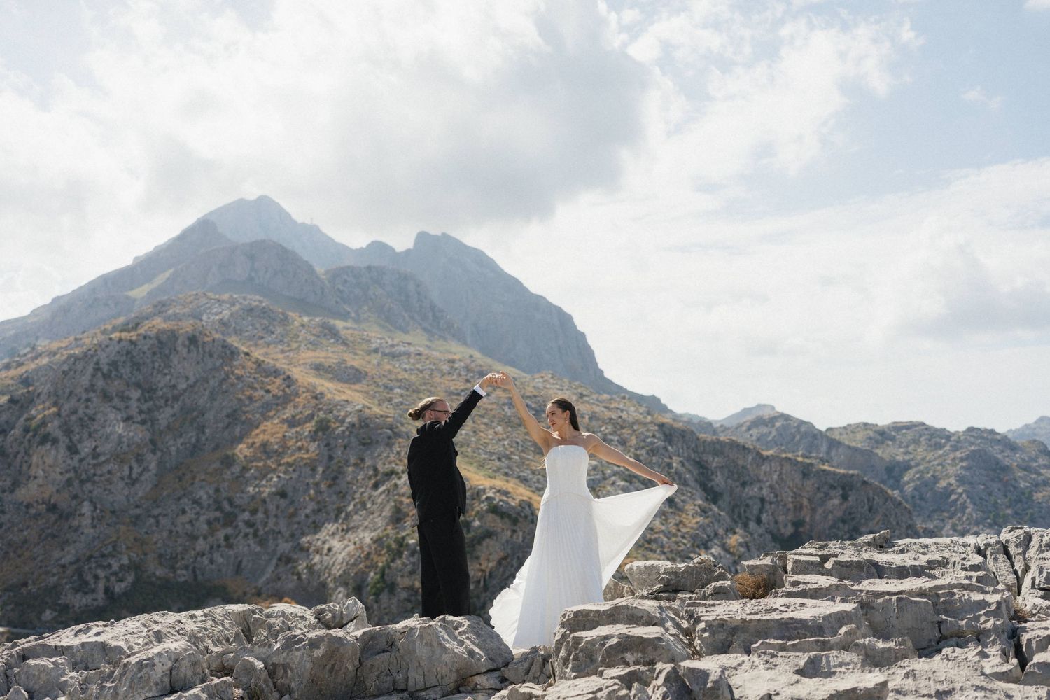 Couple in black suit and flowing white dress dance hand in hand on a rocky mountain summit with dramatic peaks and cloudy sky behind them.
