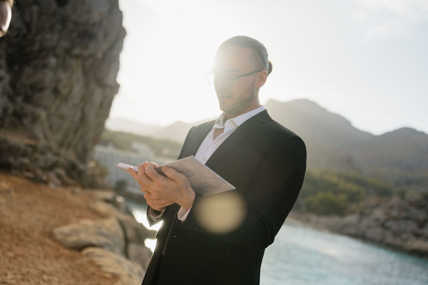 Man in a black suit and glasses reads from an open book while standing on a rocky coastline with calm sea and mountains behind him.