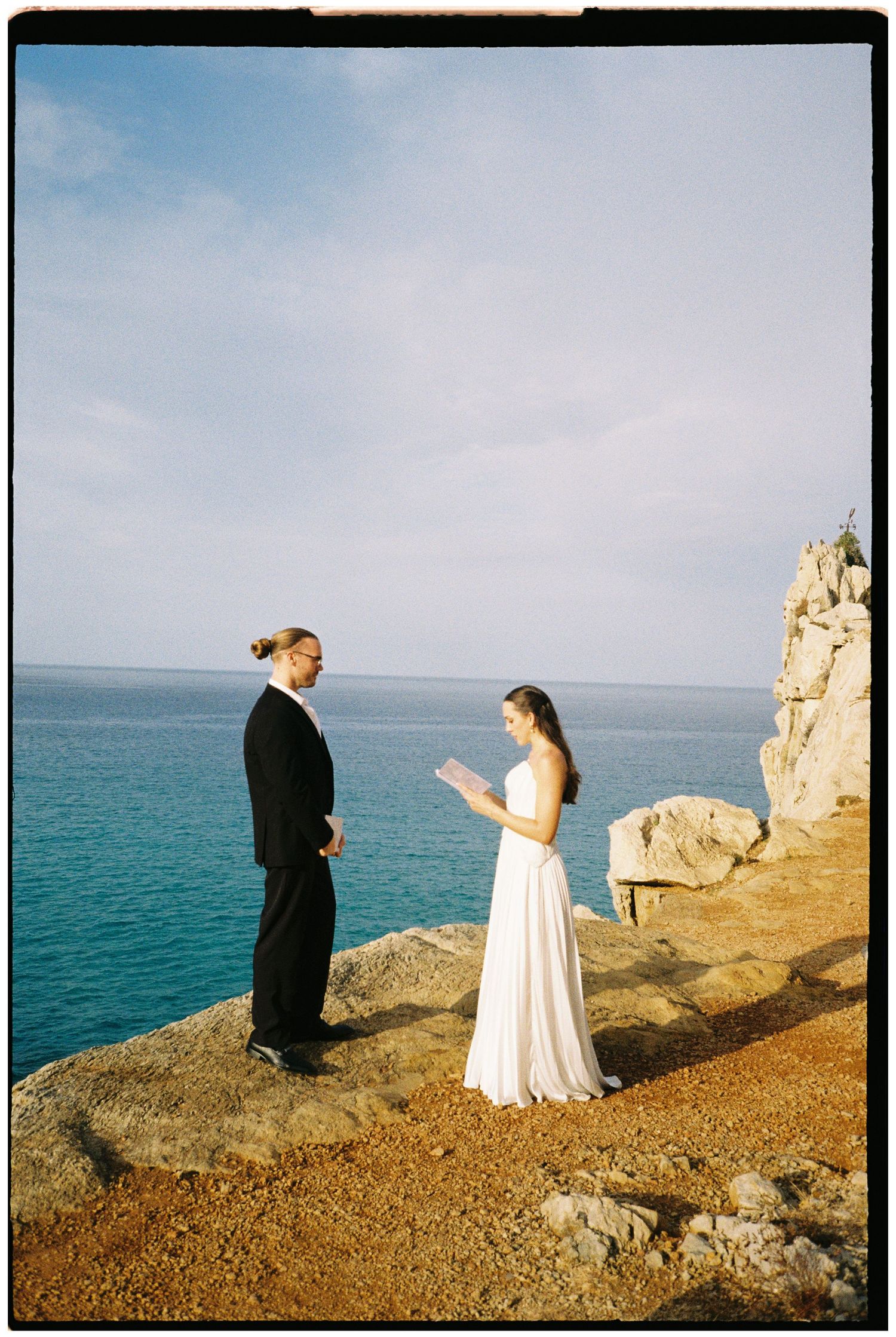 Bride in a white gown reads her vows to a groom in a black suit on a rocky cliff overlooking the turquoise sea.