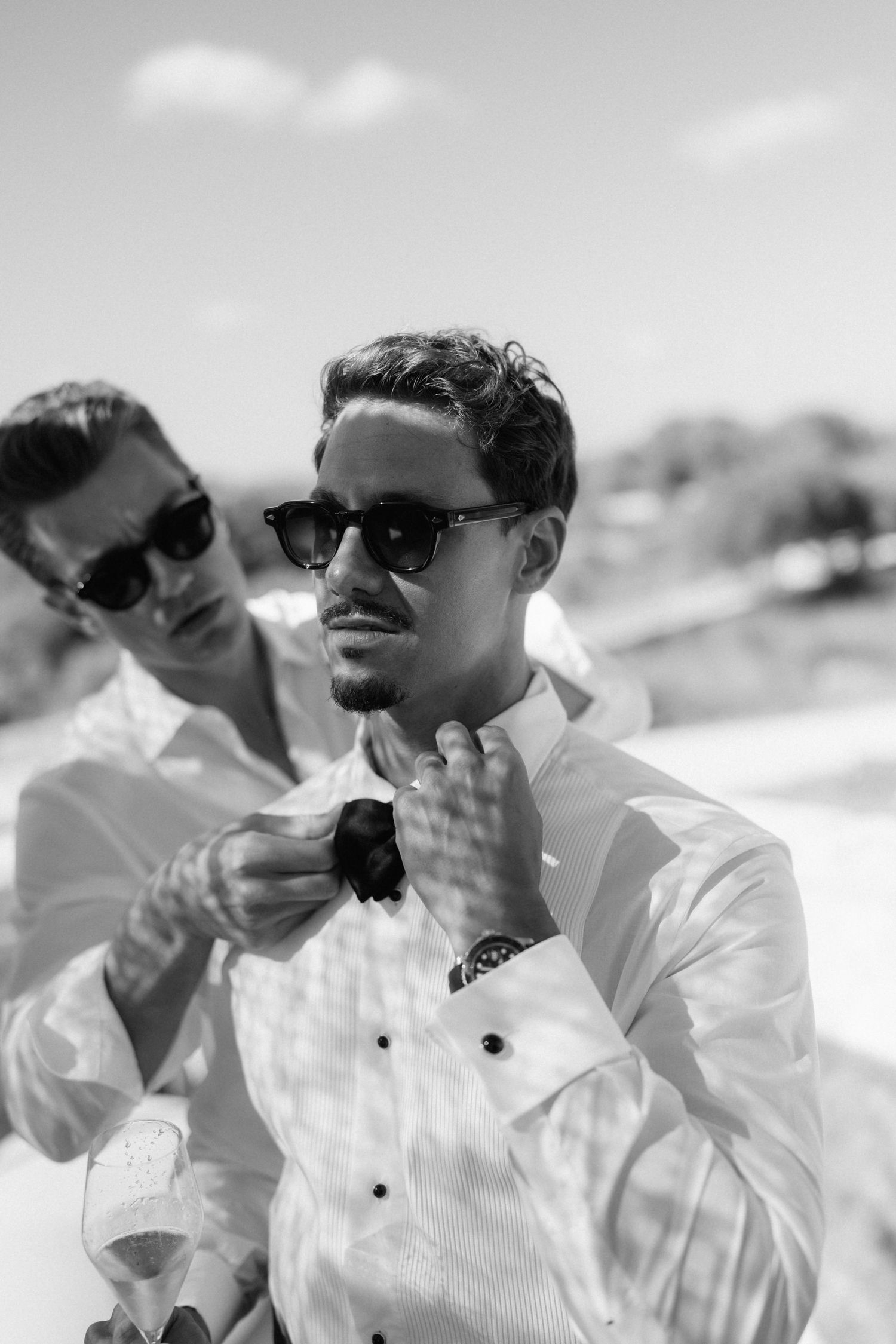Two men in sunglasses getting ready for a wedding in black and white, one adjusting a bow tie on a sunny day.