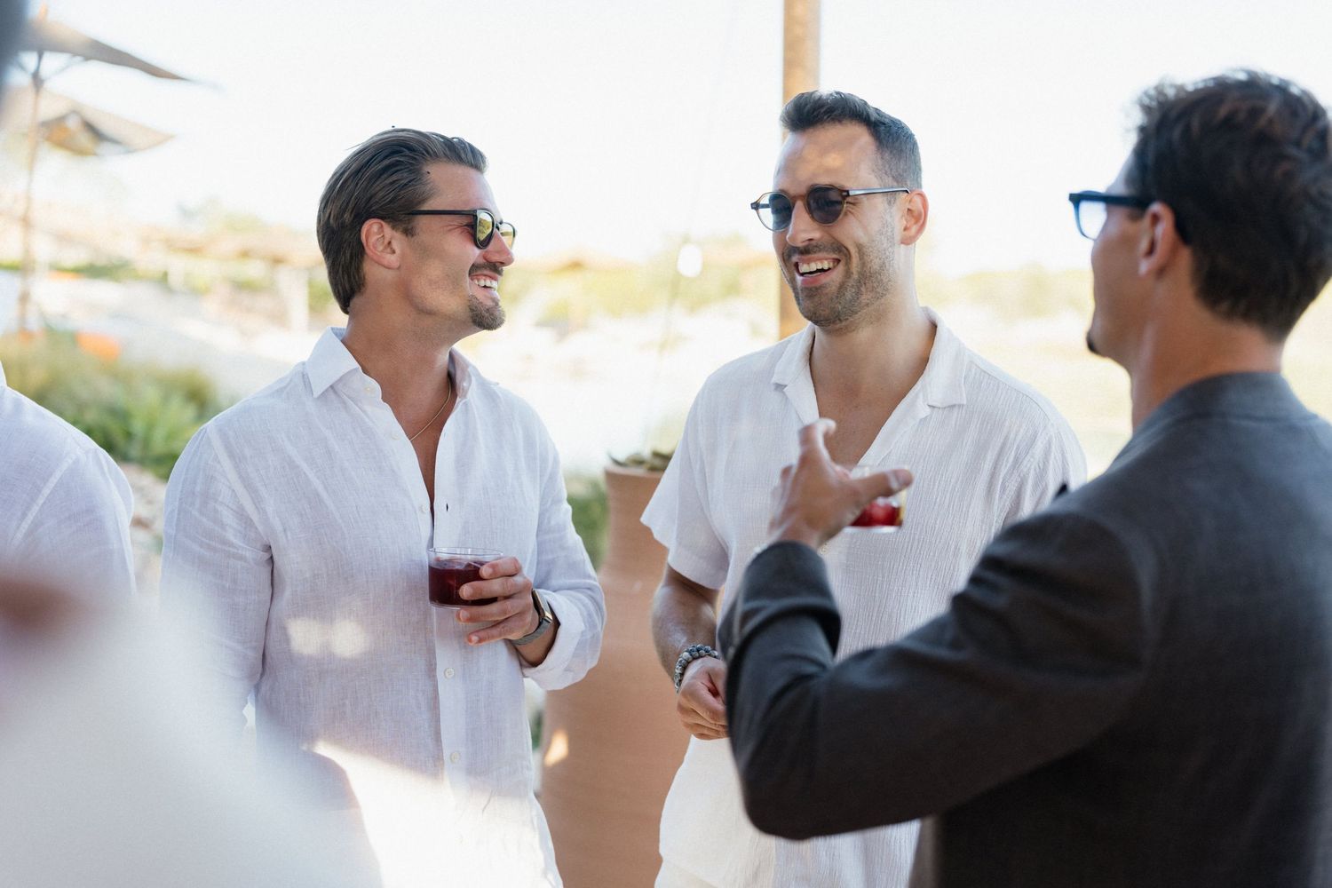 Three men in sunglasses and light linen shirts laugh and chat while holding drinks at a sunny outdoor gathering.