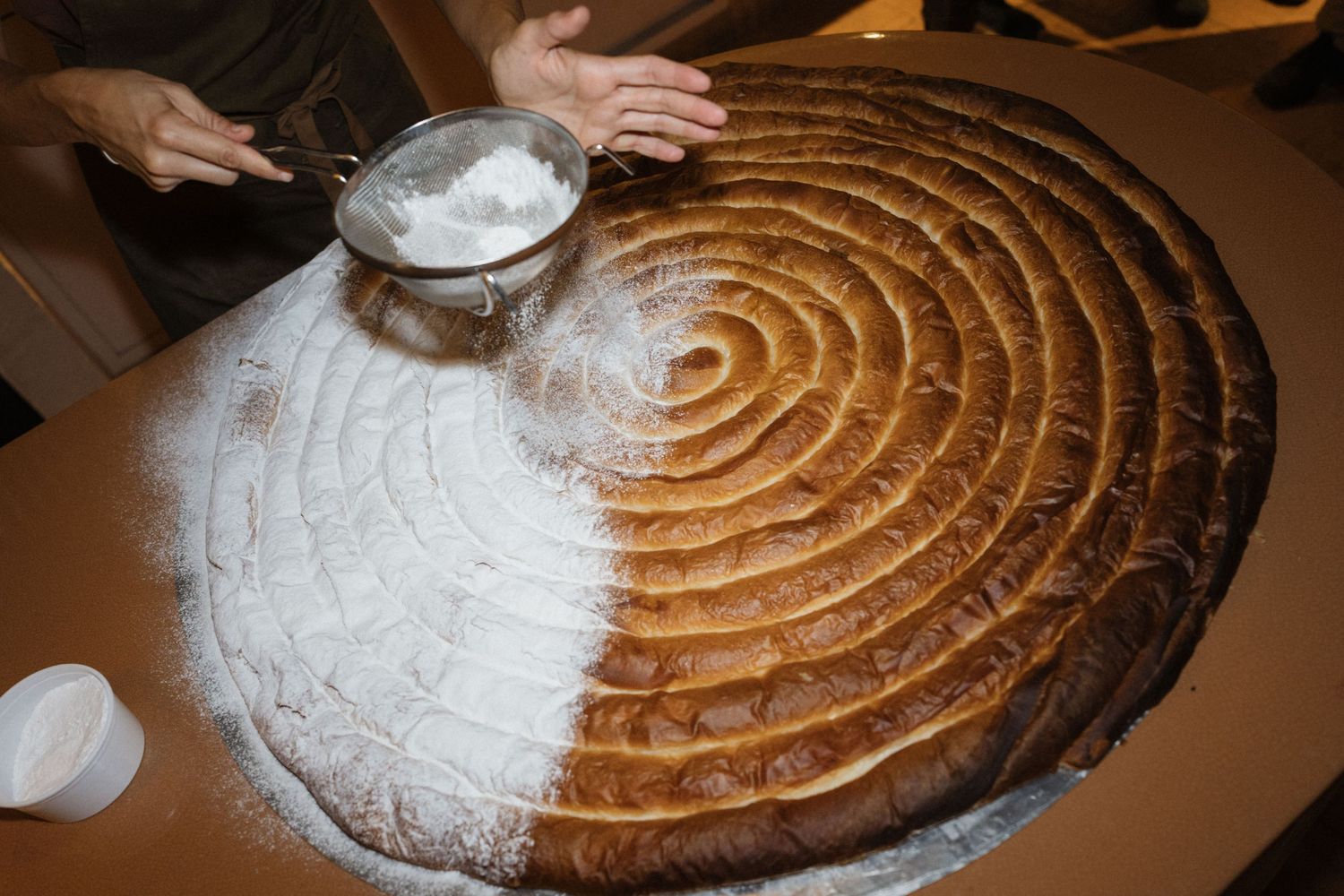 A person dusts powdered sugar through a sieve over a giant spiral-patterned pastry, half covered in sugar and half golden brown.