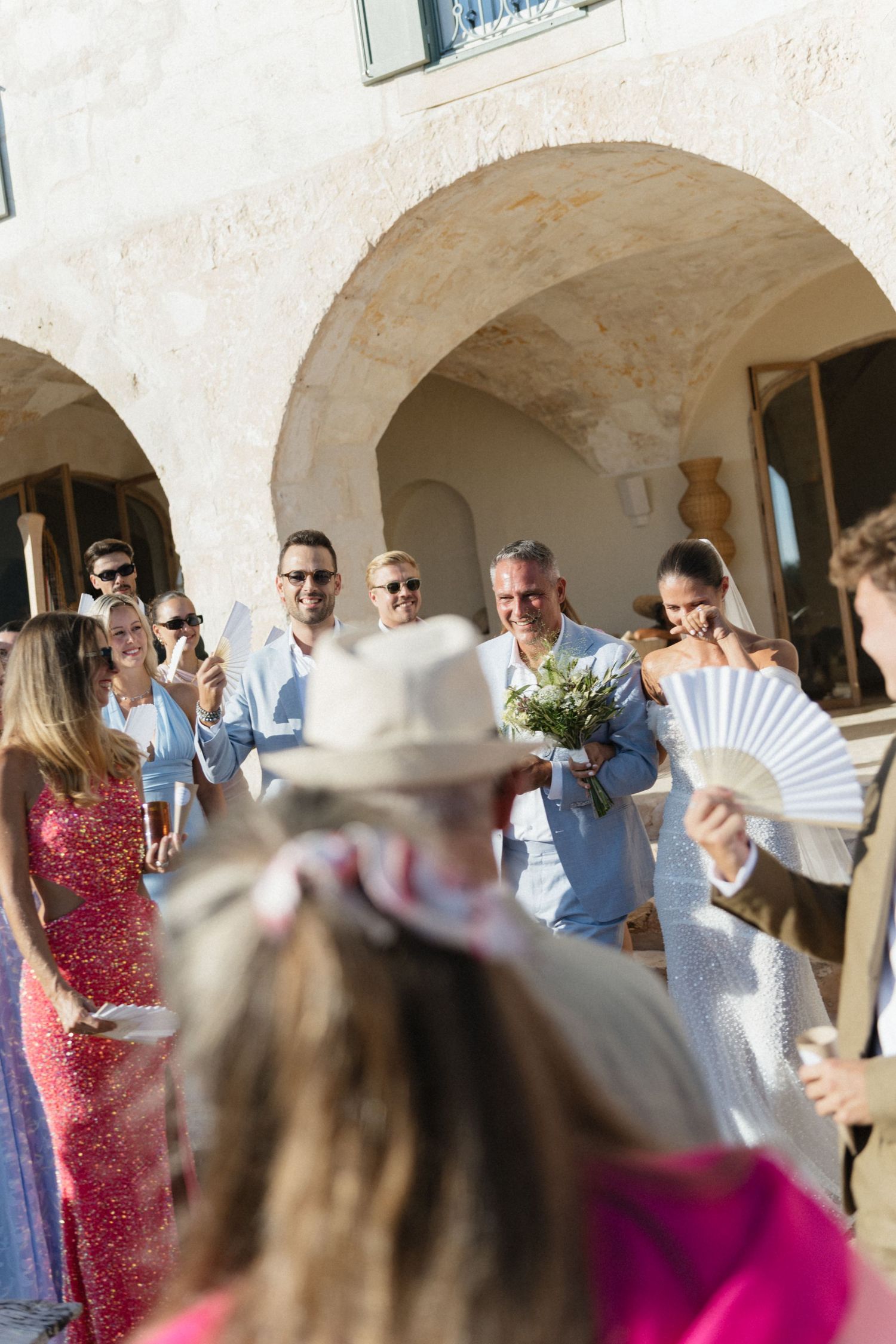 Wedding guests gather beneath a stone archway as a groom holding a bouquet walks with a bride, while a guest holds a fan.