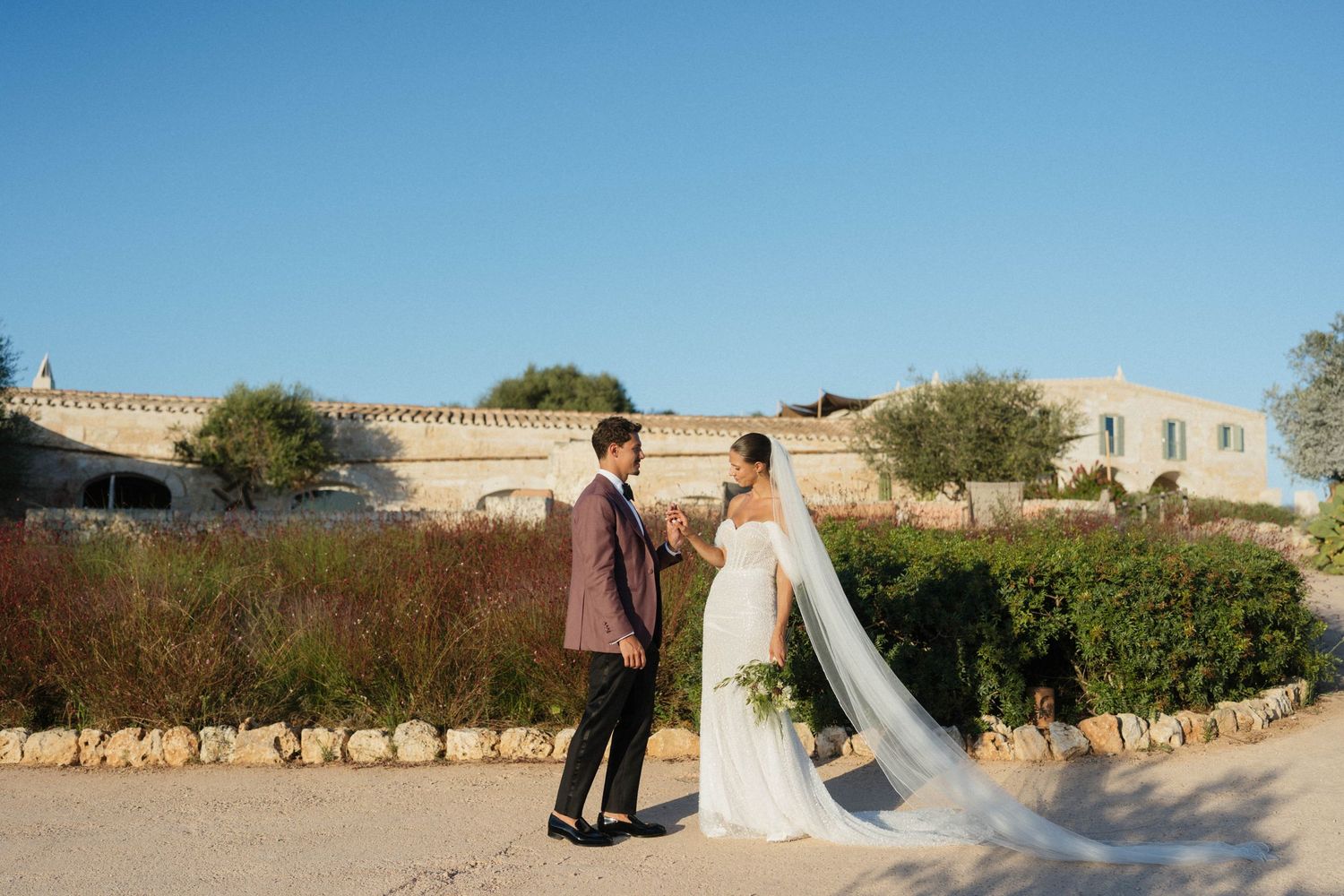 A bride and groom in a burgundy jacket share a quiet moment in a sunlit Mediterranean garden with a stone villa behind.