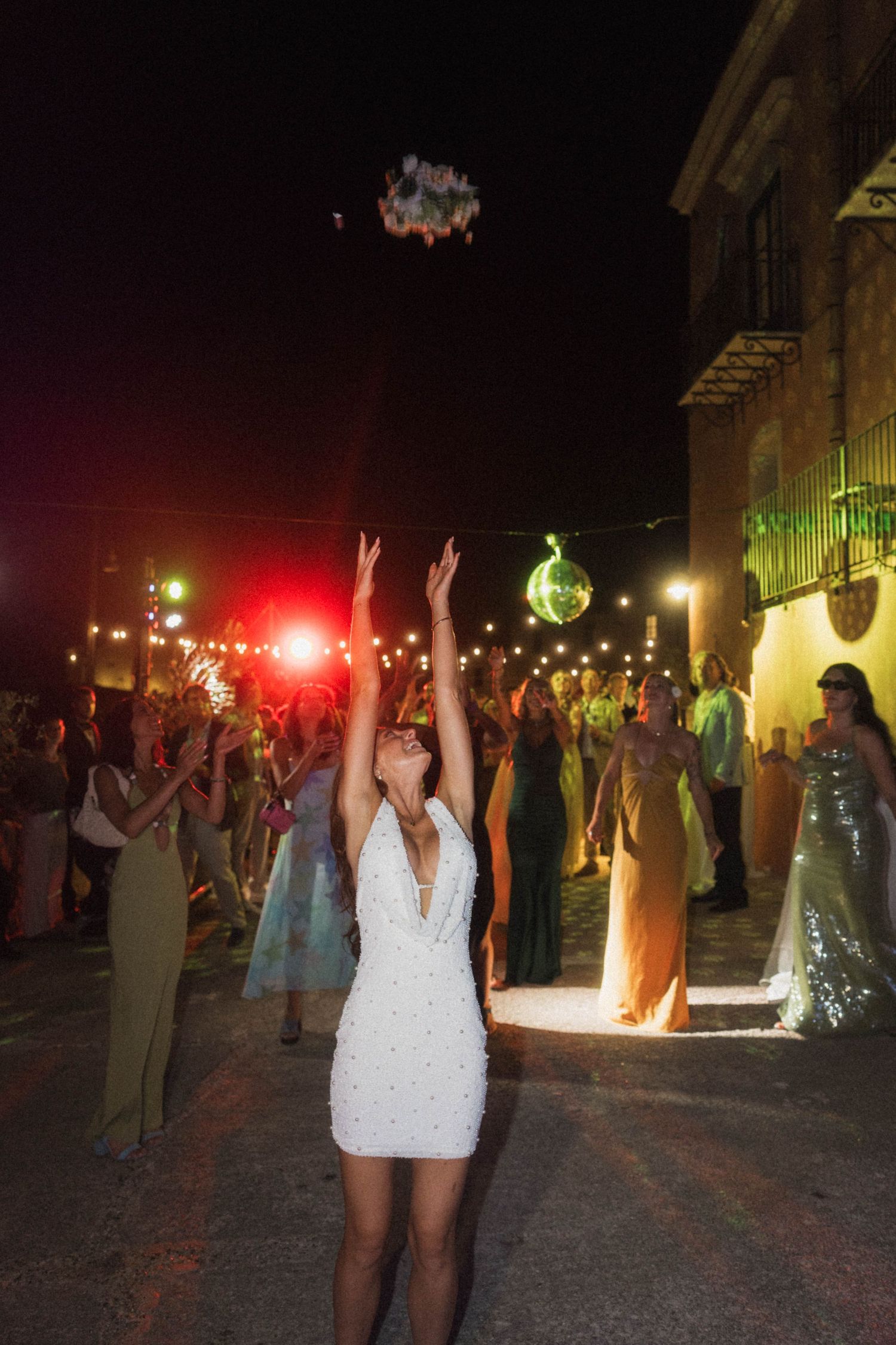 Bride in white halter mini dress throwing bouquet to guests at outdoor nighttime wedding.