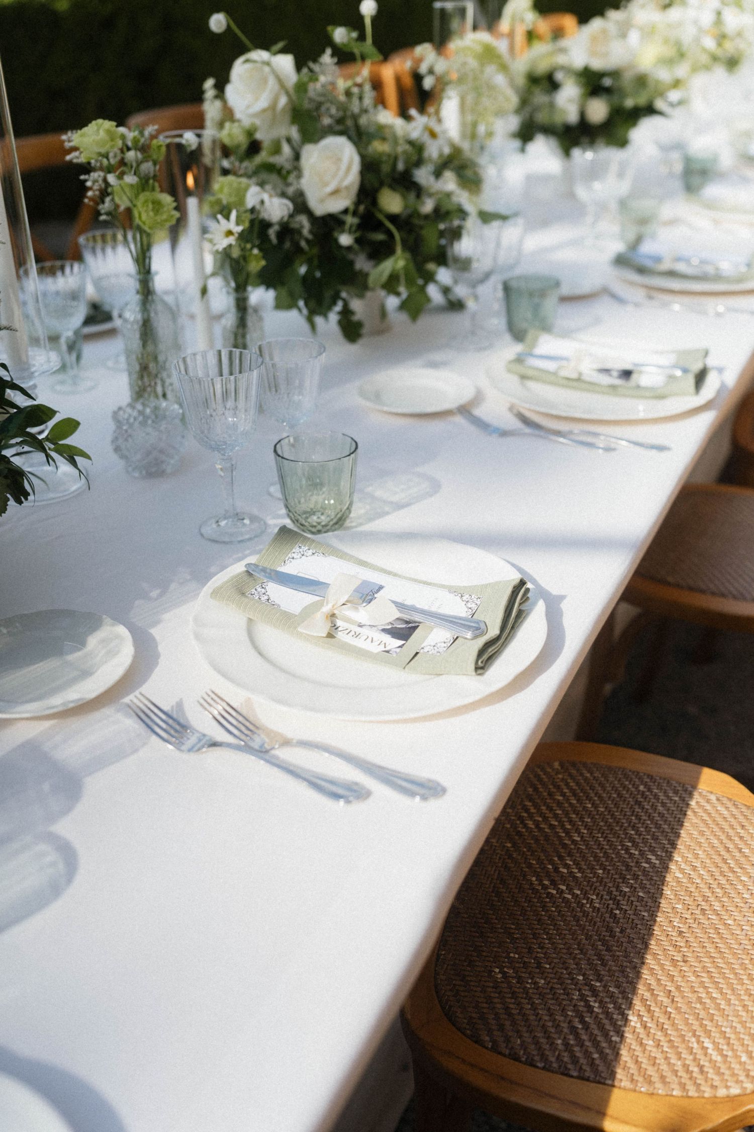 Elegant wedding table setting with white plates, green glassware, silver cutlery, and white floral centerpieces.