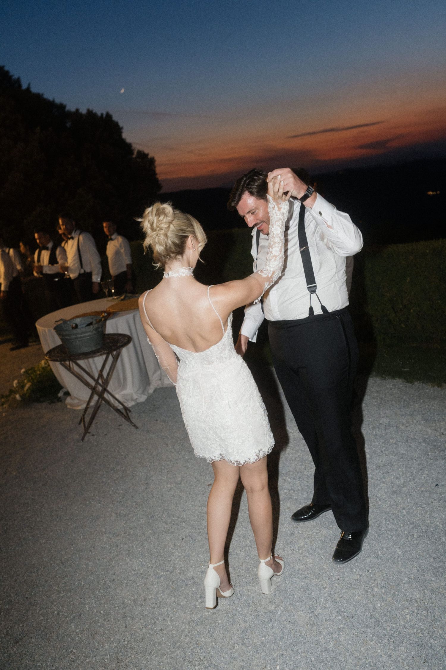 Bride and groom dancing outdoors under a colorful twilight sky during their wedding reception.