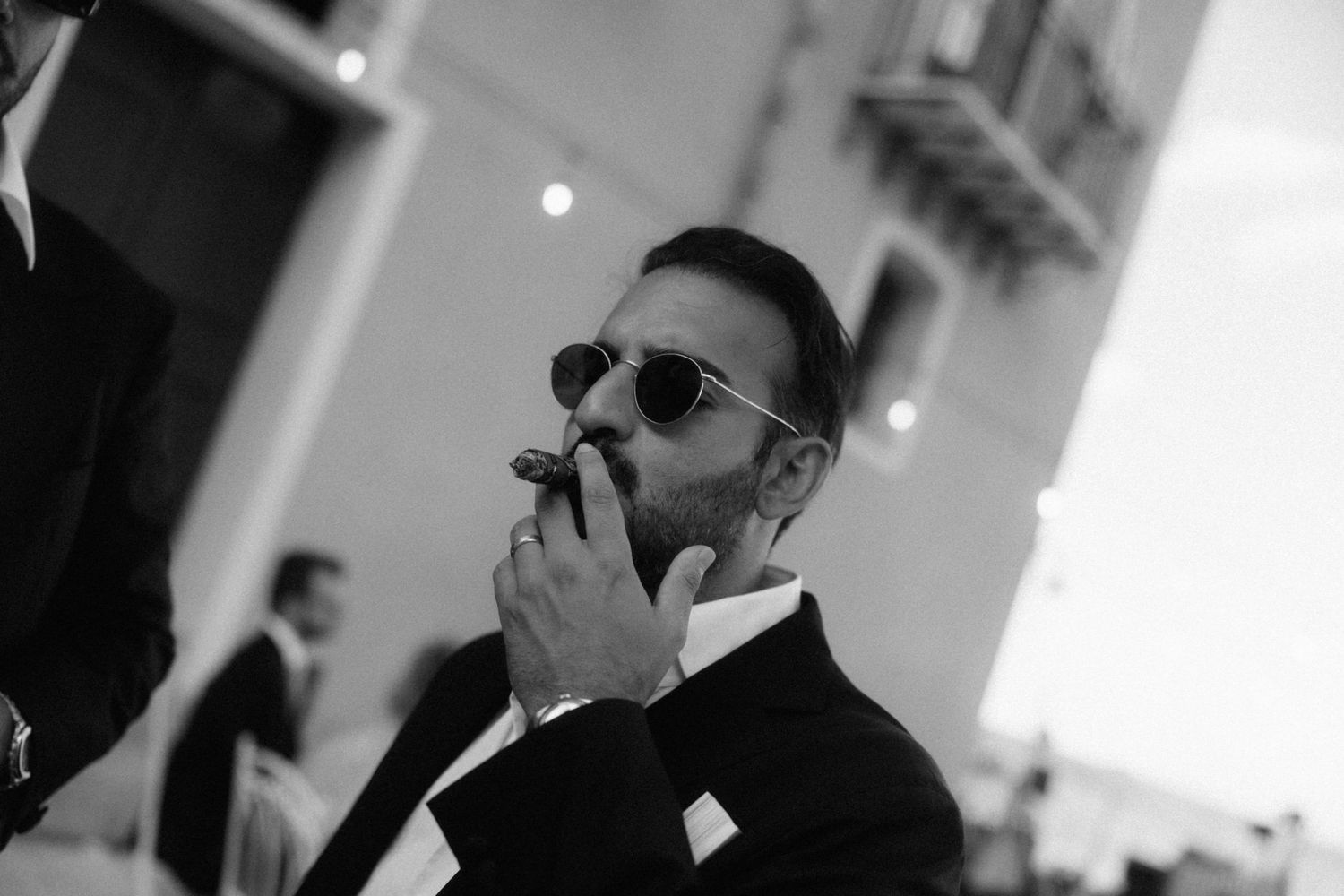 Stylish bearded groom in black suit and sunglasses lighting a cigar at an outdoor wedding reception.