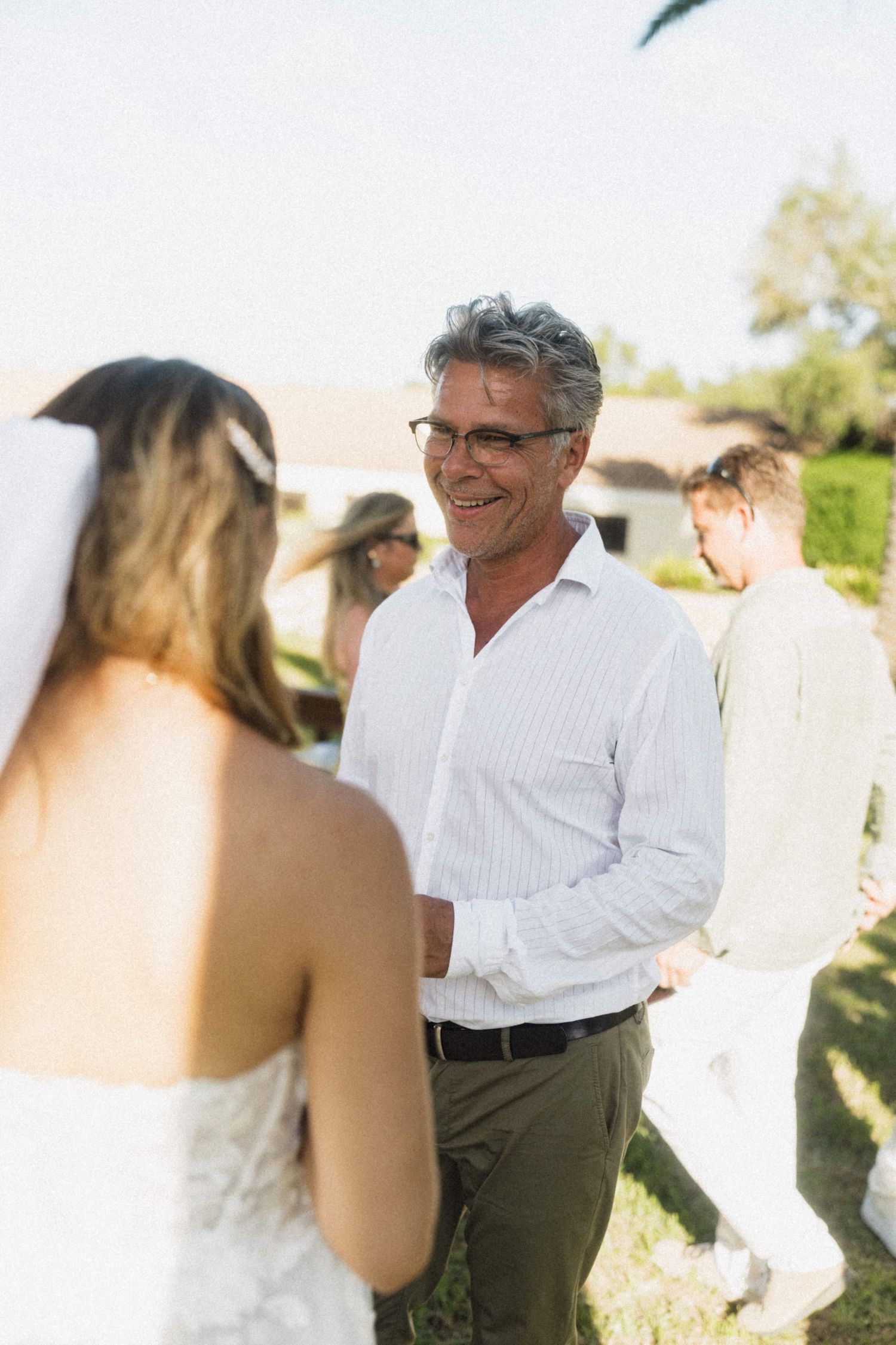 Smiling older man with glasses greets bride in white lace gown at sunny outdoor wedding.