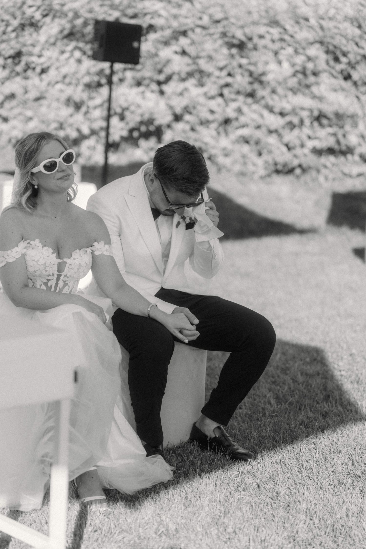 Black and white photo of bride and groom sitting outdoors at wedding ceremony, groom emotional wiping tears.