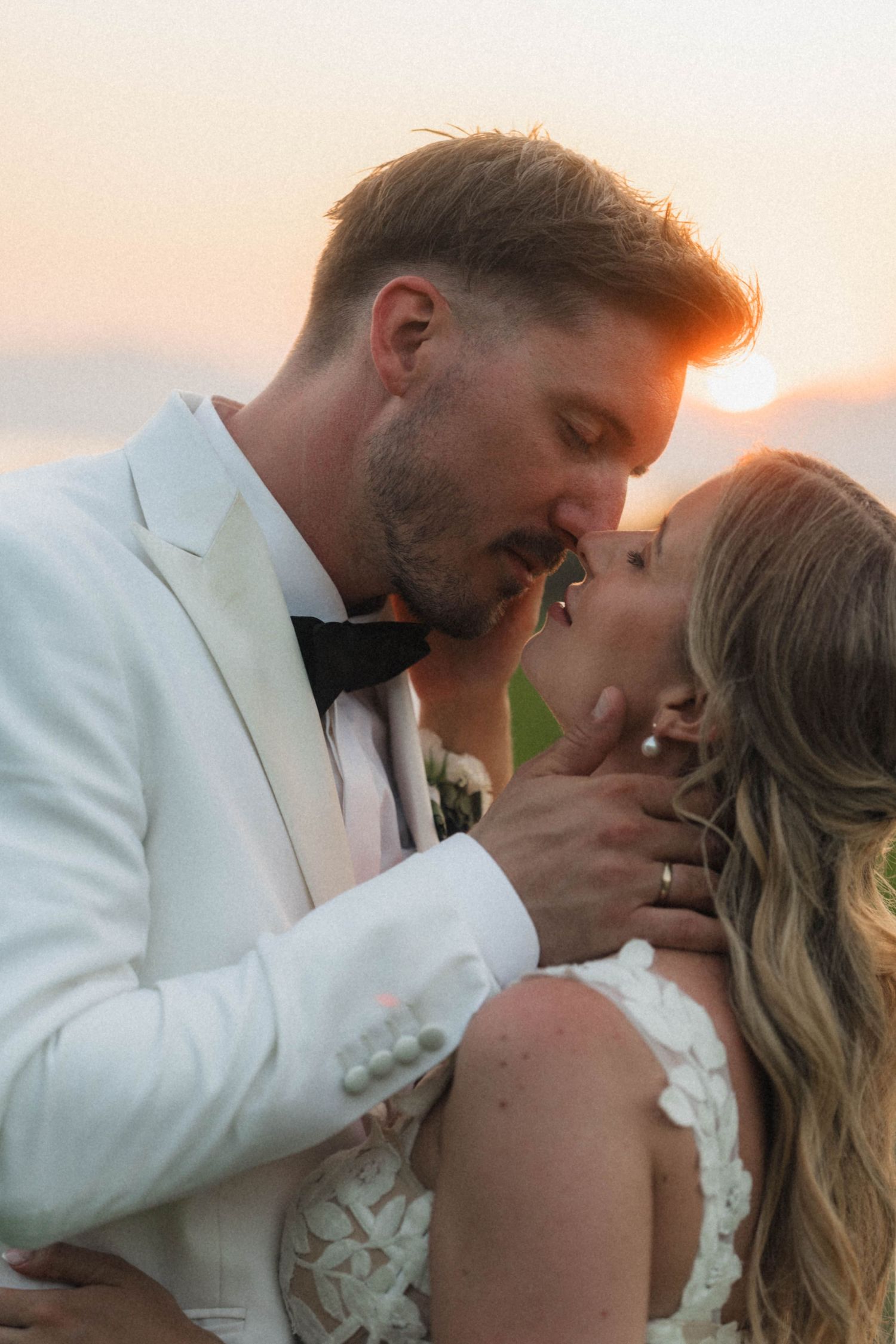 Bride and groom sharing romantic kiss at golden sunset during outdoor wedding portraits.