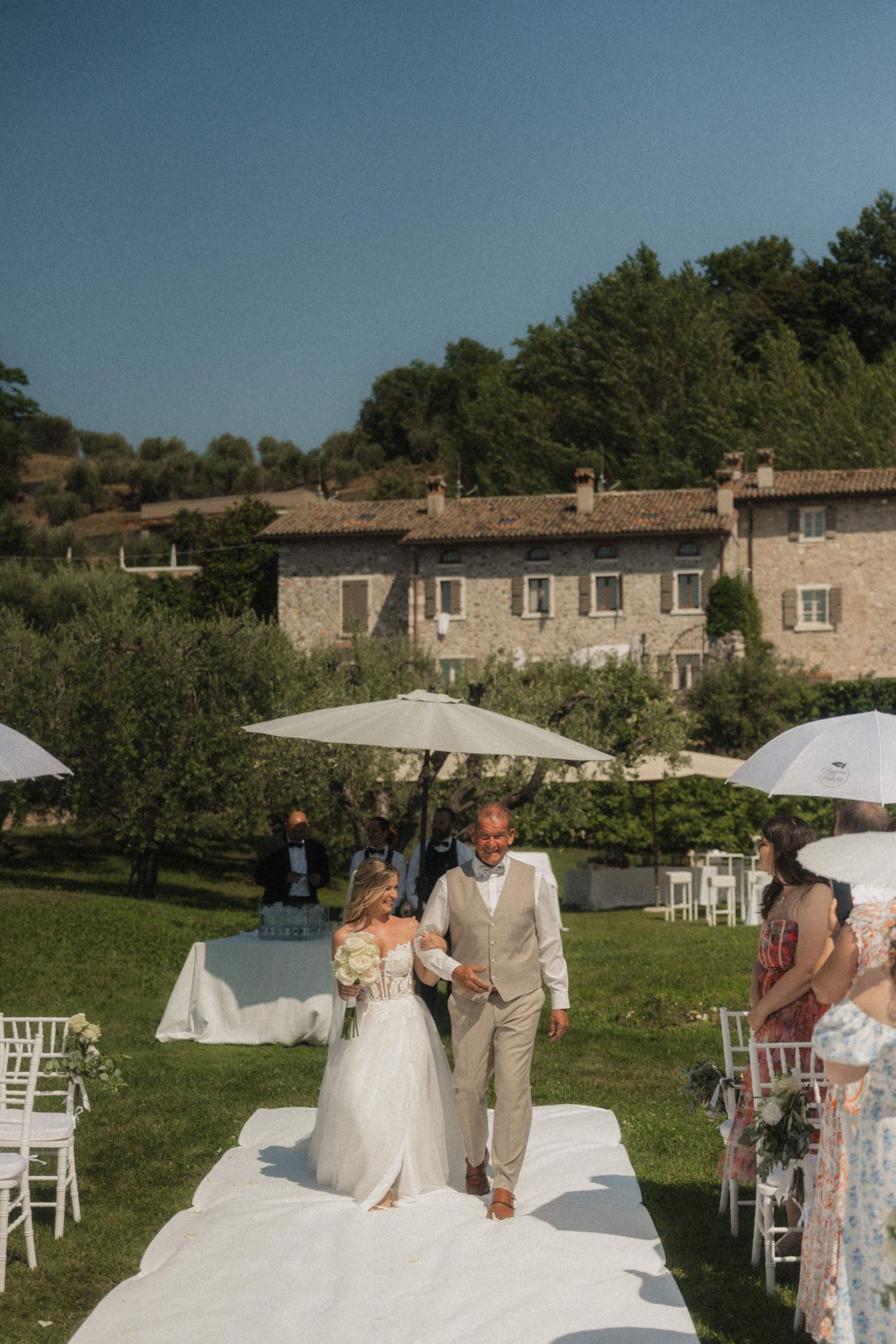 Bride and groom walk down white aisle at outdoor wedding ceremony with stone villa backdrop in Italy.