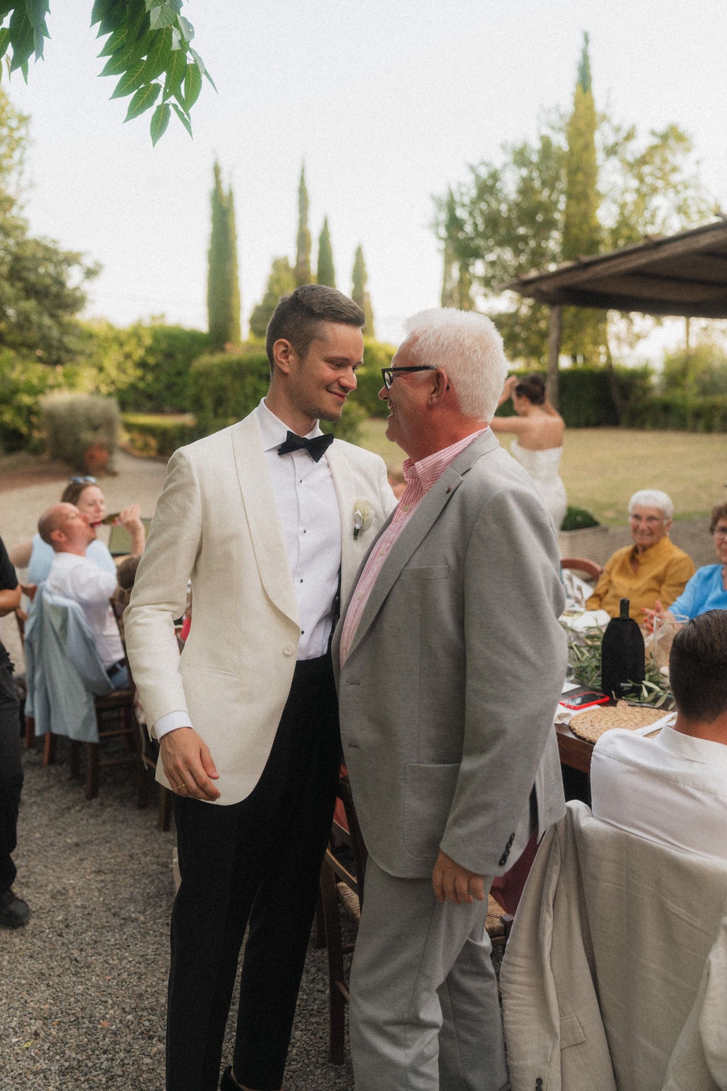 Groom in white tuxedo jacket sharing a moment with older gentleman in gray suit at wedding reception.