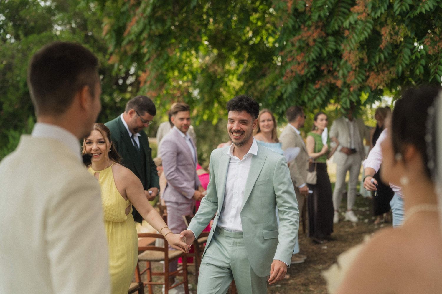 Groom in mint green suit smiling at outdoor wedding ceremony surrounded by guests under lush green trees.