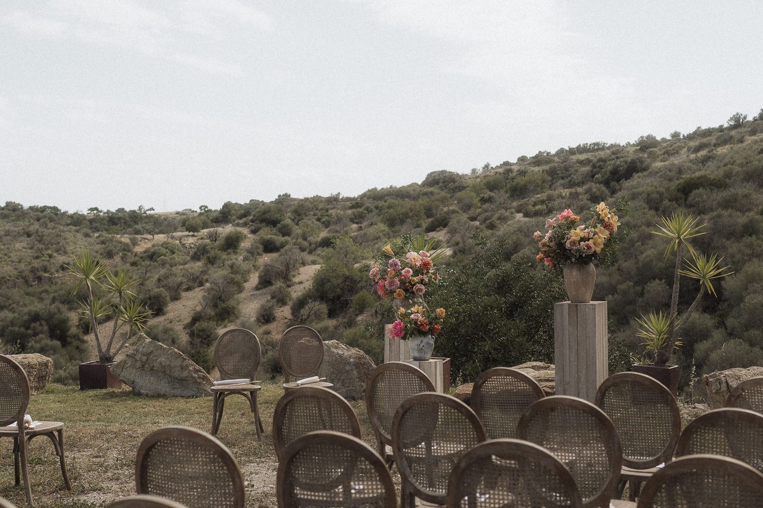 Outdoor wedding ceremony setup with wicker chairs and floral arrangements on wooden pedestals against a scenic hillside backdrop.