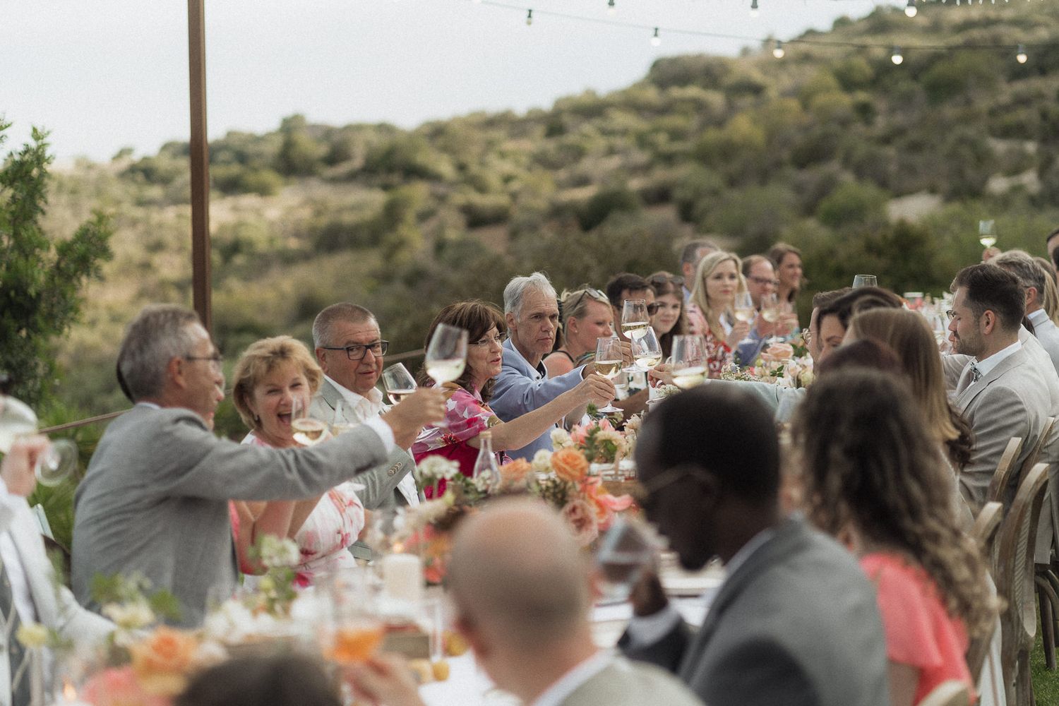 Guests gathered at an outdoor wedding reception dinner table toasting amid lush green hillside scenery.