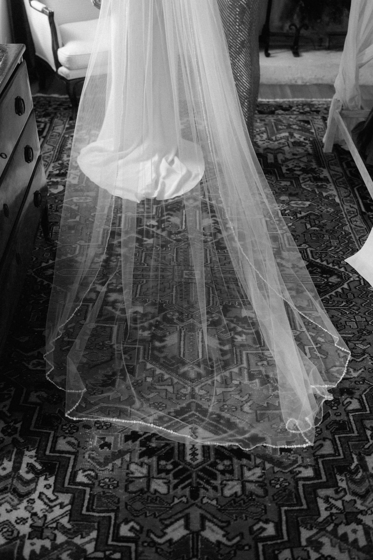 Close-up black and white photo of flowing bridal veil and gown train spread across ornate patterned rug.