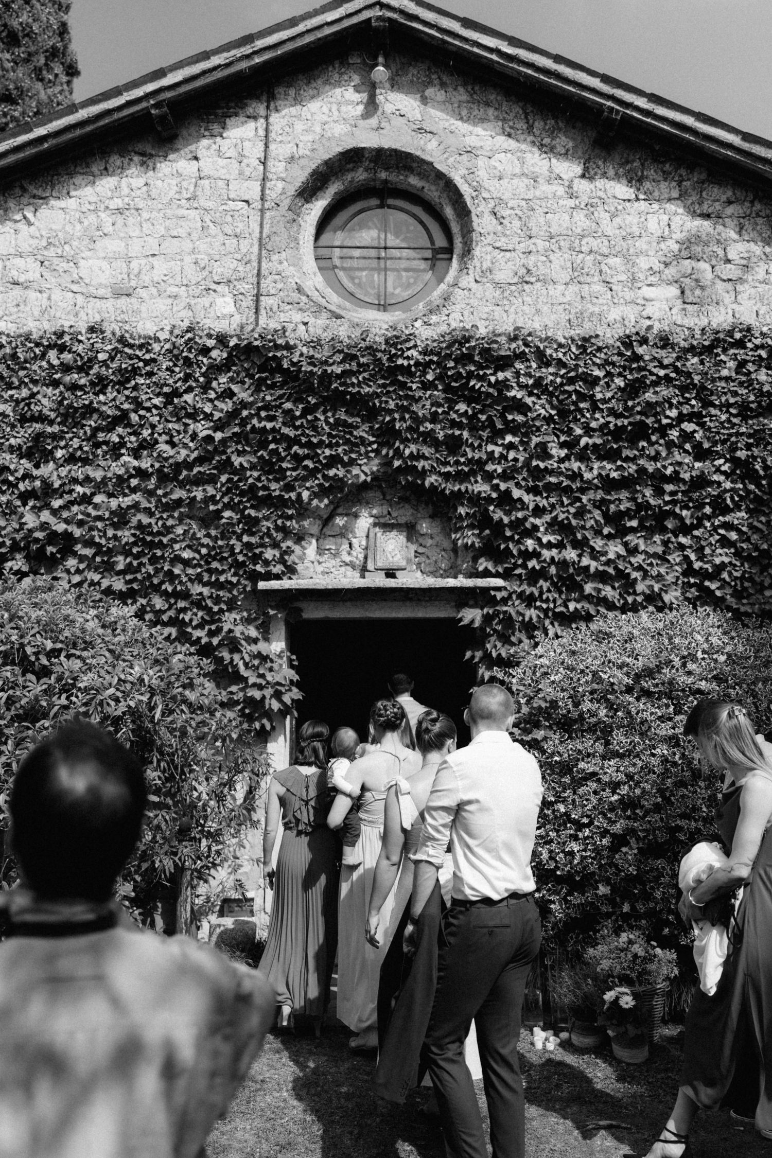 Wedding guests entering an ivy-covered stone chapel with a round window for a ceremony.