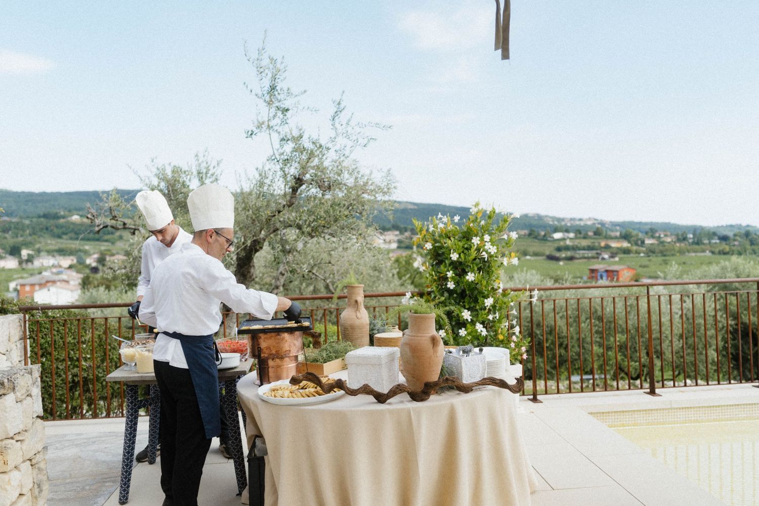 Chef preparing food at an outdoor terrace buffet station with scenic countryside views and olive trees.