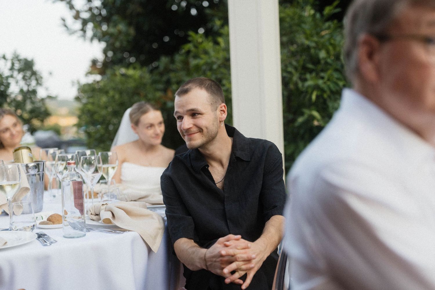 Man in black shirt smiling at an elegant outdoor wedding reception dinner table with guests.