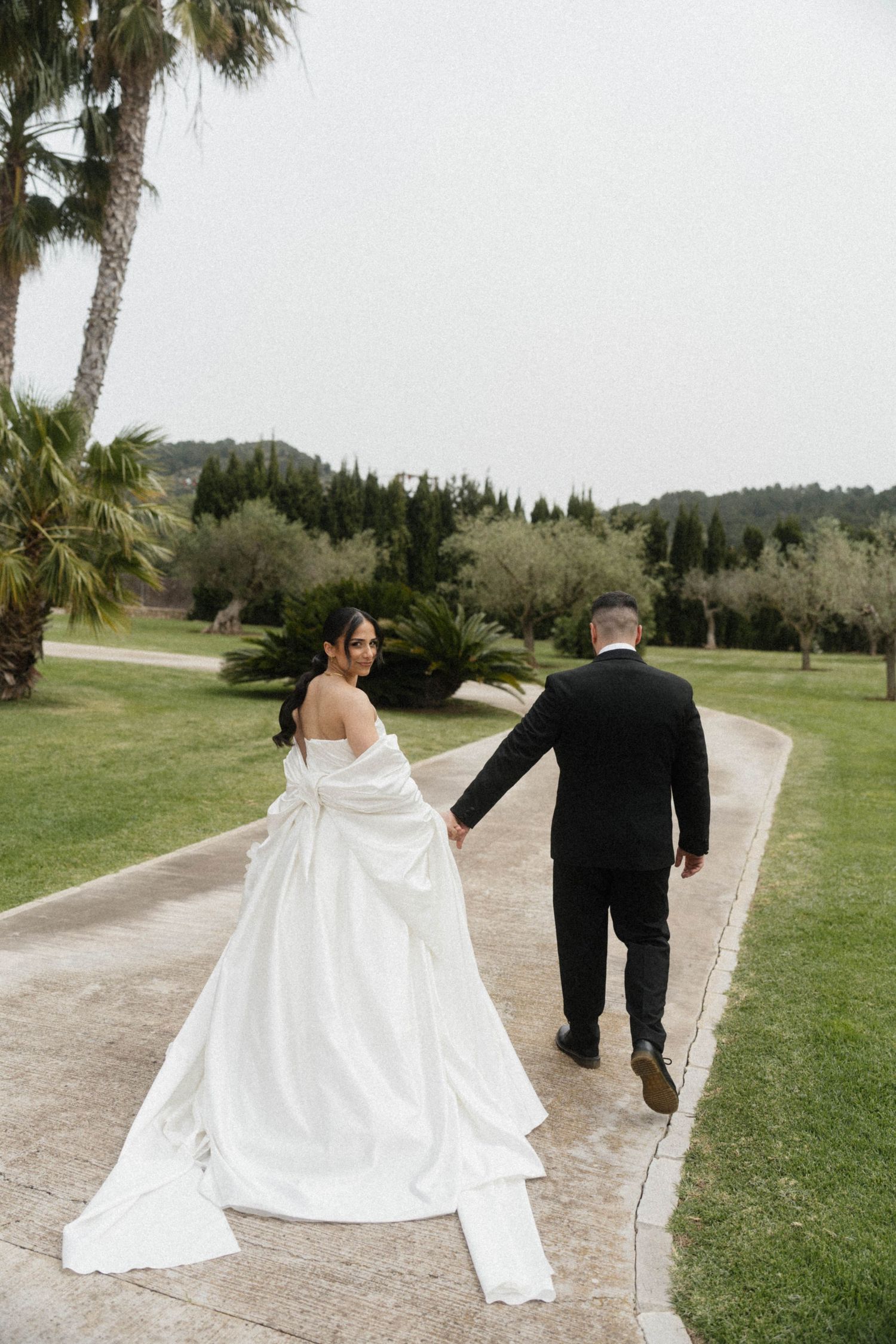 Bride in white gown and groom in black suit walk hand in hand along a garden path at an outdoor wedding.
