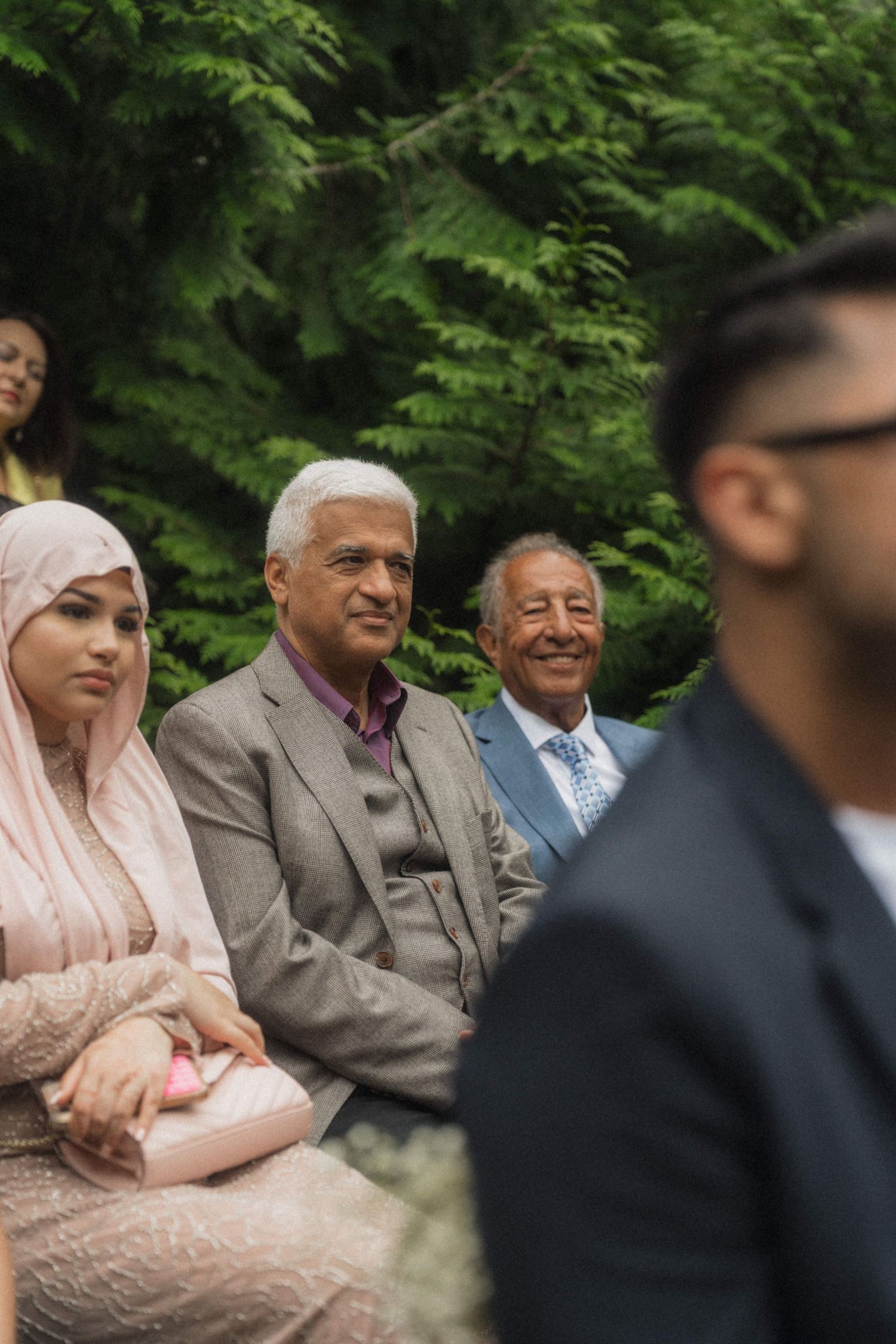 Smiling wedding guests seated outdoors beneath green pine trees watching an outdoor wedding ceremony.