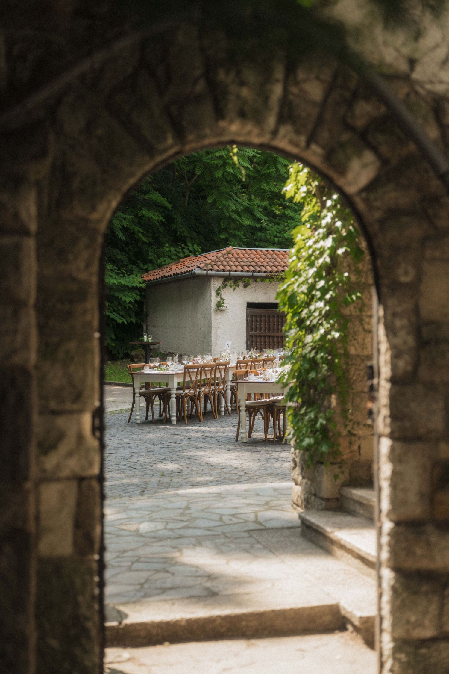 Stone archway frames an outdoor wedding reception dining area with long tables set amid lush greenery.