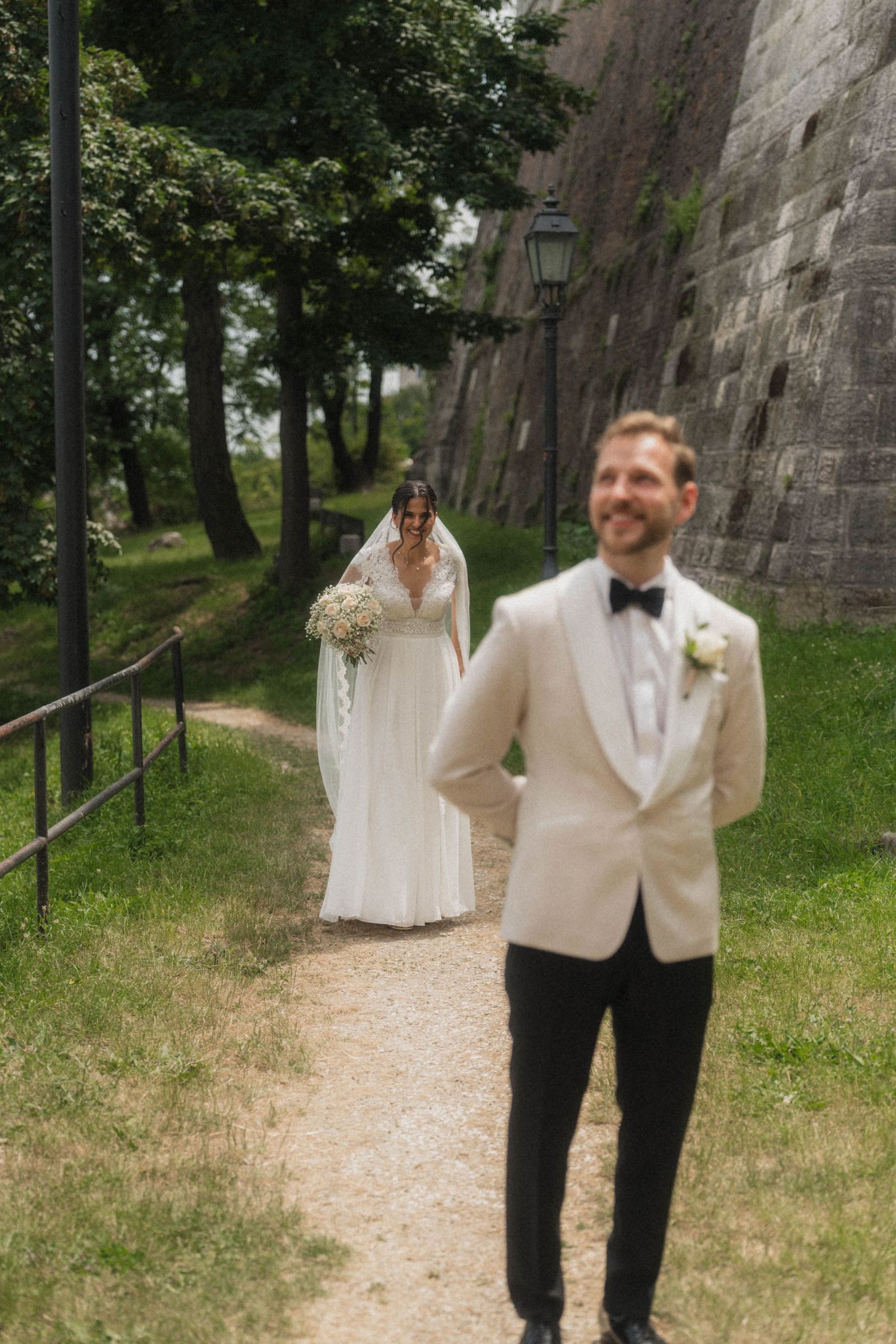 Bride in white gown with veil approaches smiling groom in cream tuxedo on a garden path near stone walls.