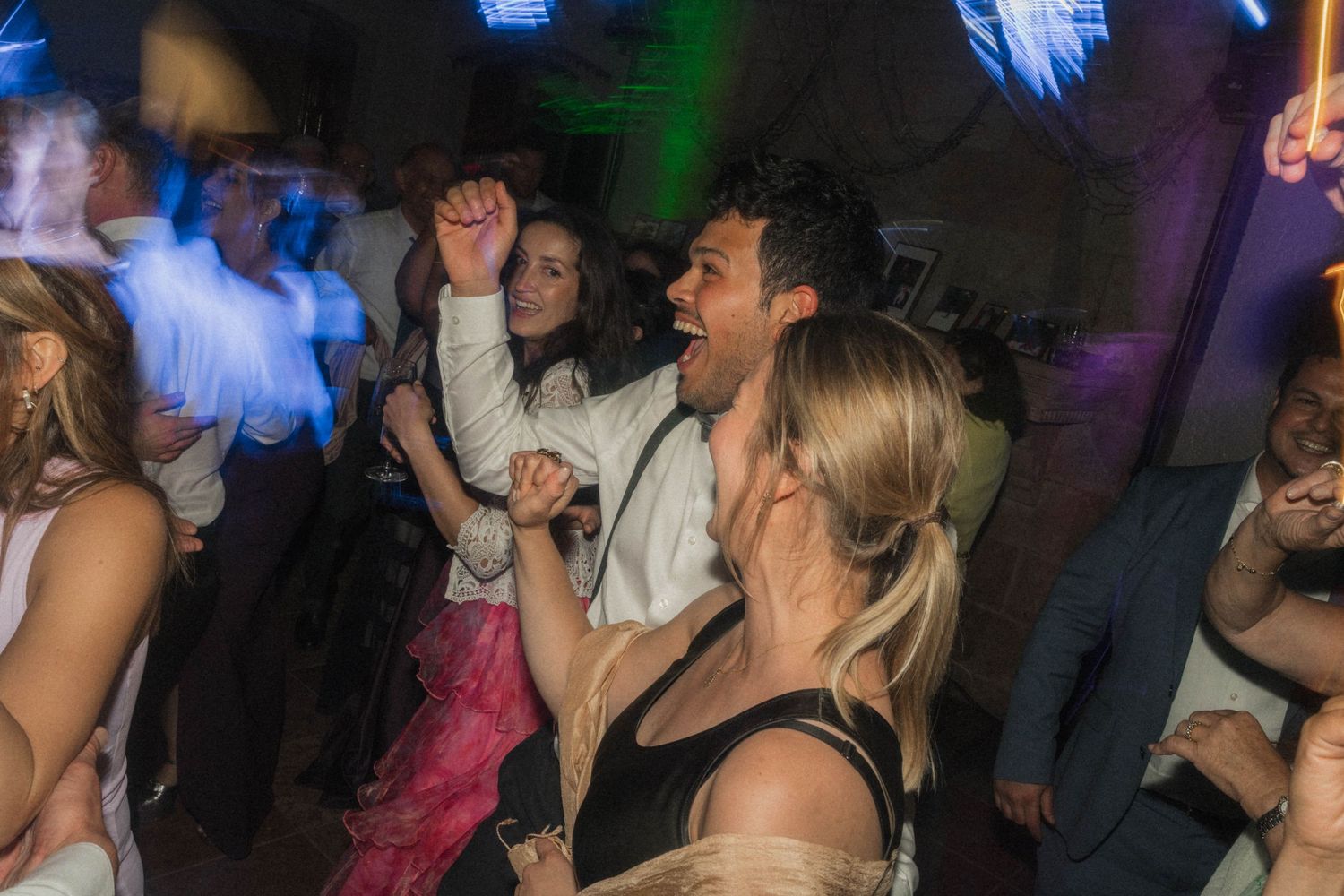 Energetic wedding guests dancing together on a colorfully lit reception dance floor.